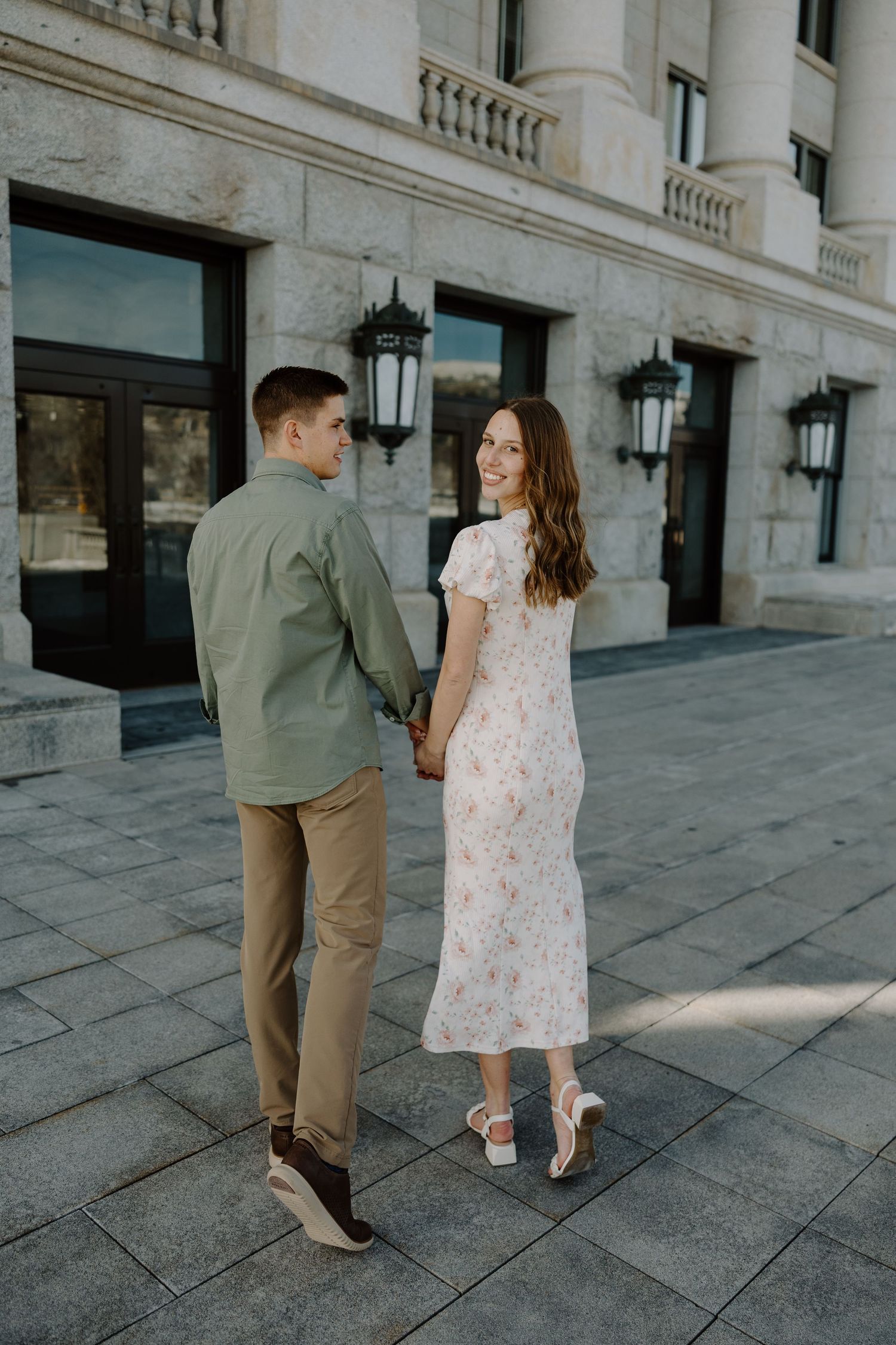 Couple holding hands walking near elegant stone building, woman in floral midi dress smiling back at camera.