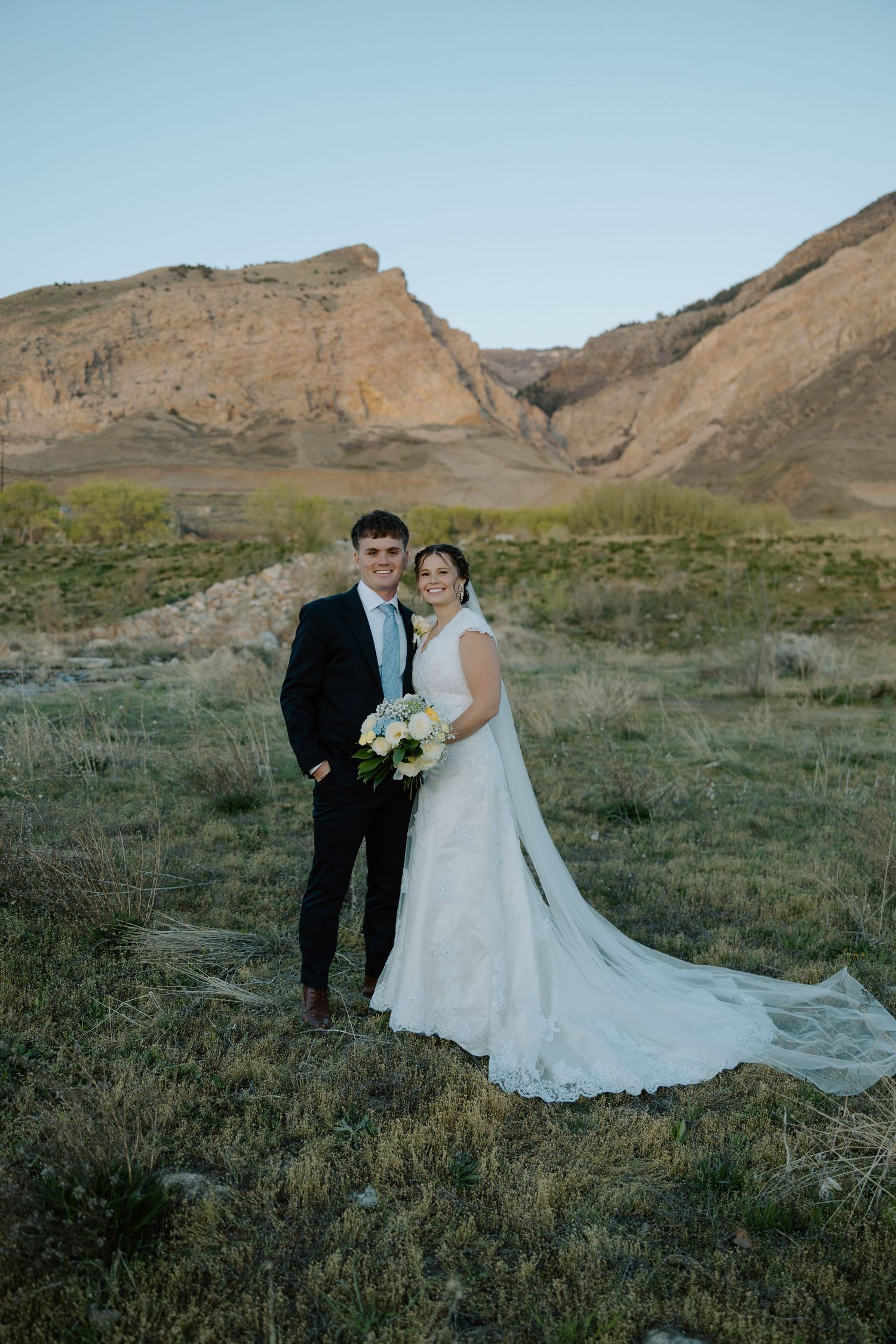 Bride and groom pose together in a scenic mountain landscape with golden rocky cliffs and open fields at sunset.
