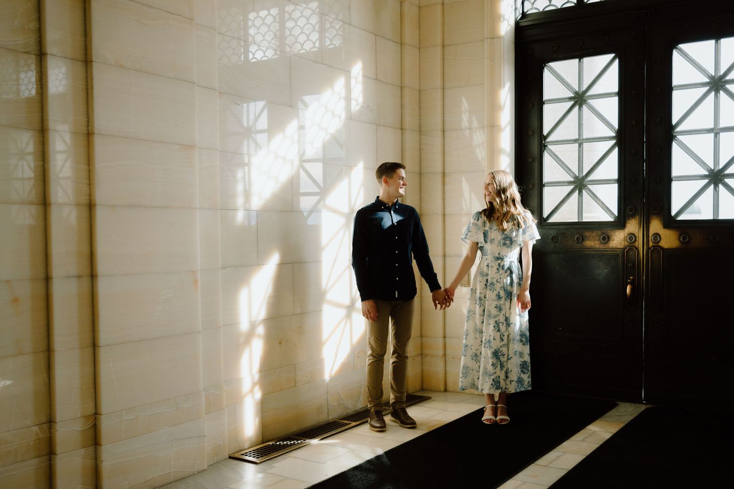 Couple holding hands in sunlit marble hallway near ornate black door, man in suit and woman in floral dress.