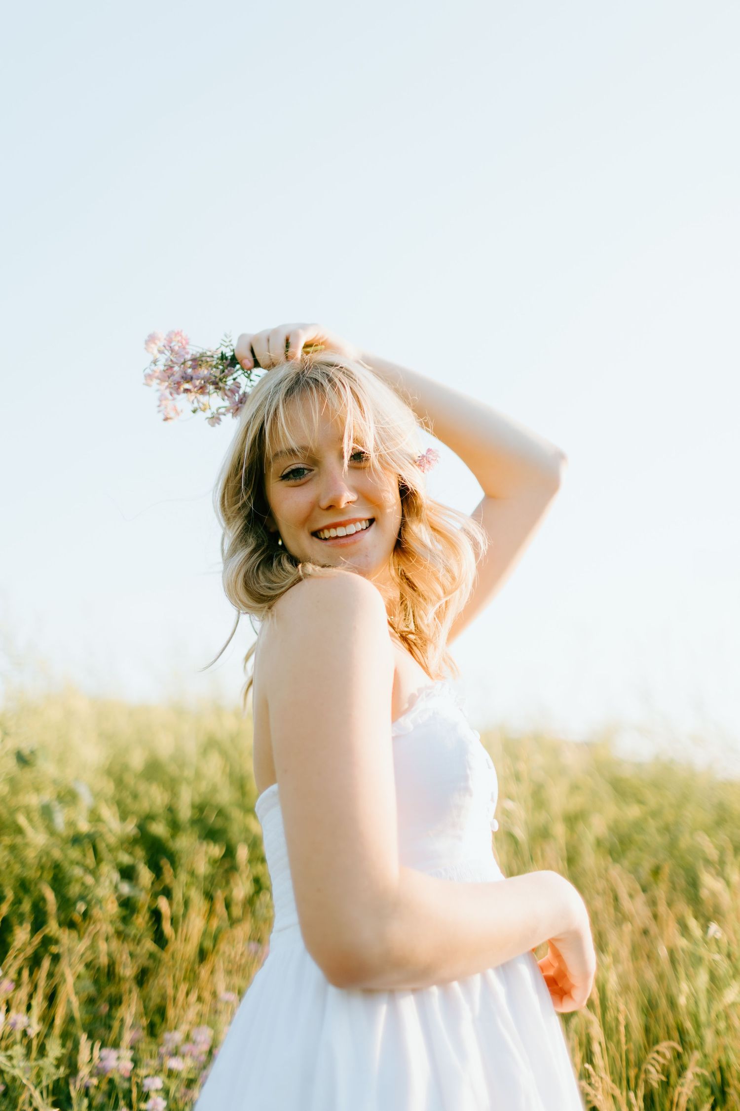 Smiling blonde woman in white dress holding wildflower crown in sunny field.