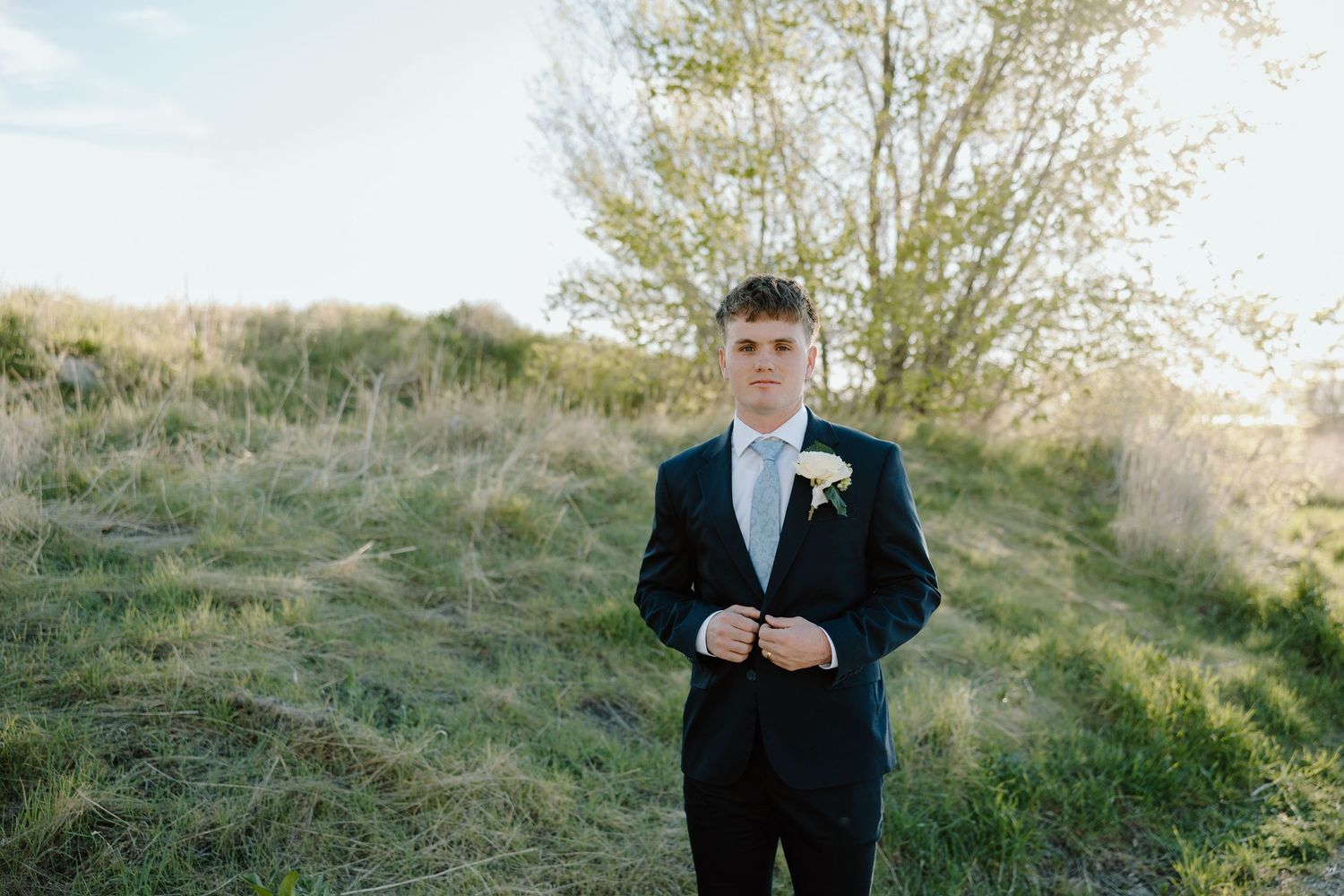 Handsome groom in black suit with white boutonniere standing in sunny green meadow.