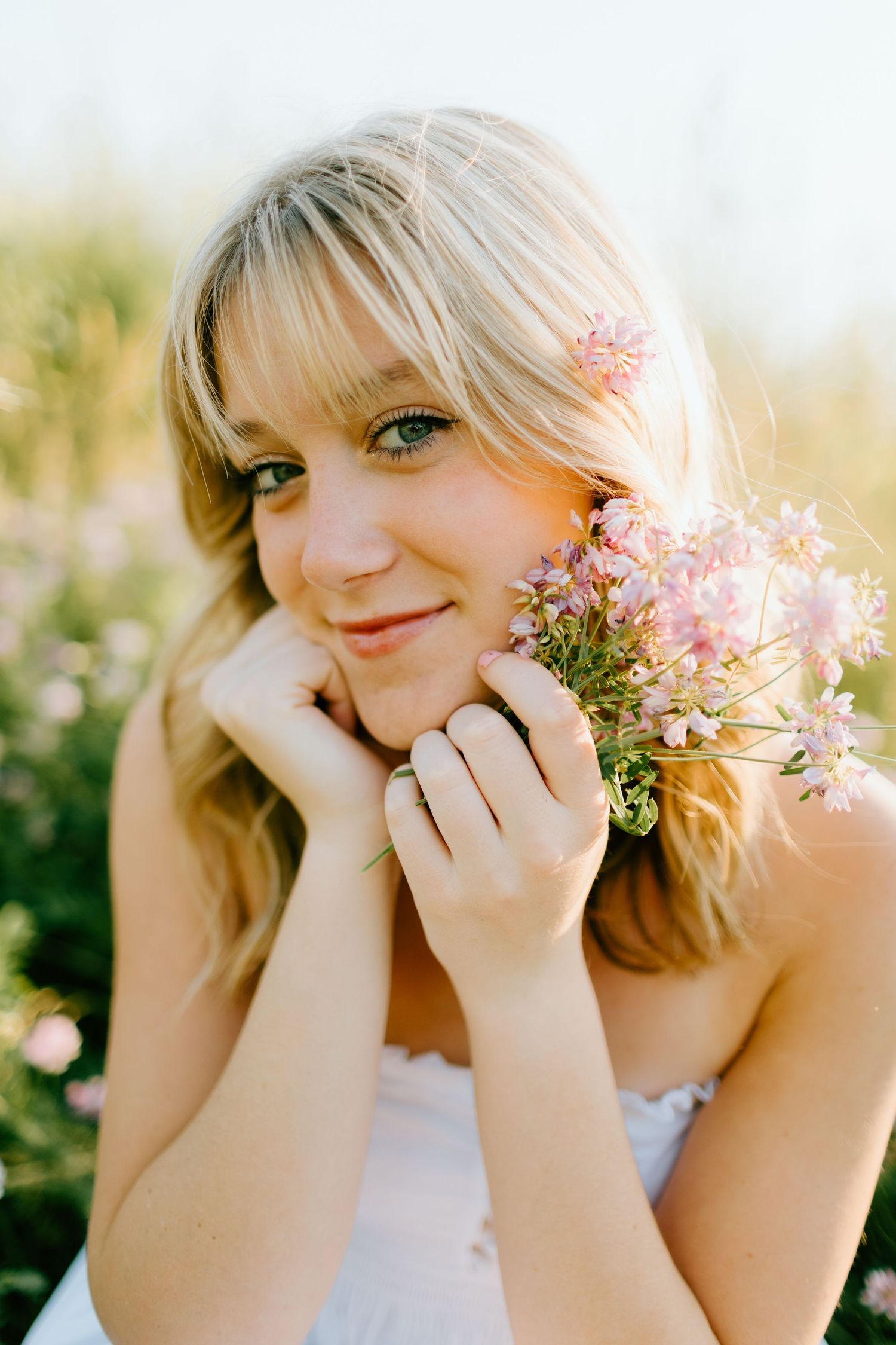 Smiling blonde woman holding pink wildflowers in a sunlit garden, wearing a white dress with a flower in her hair.