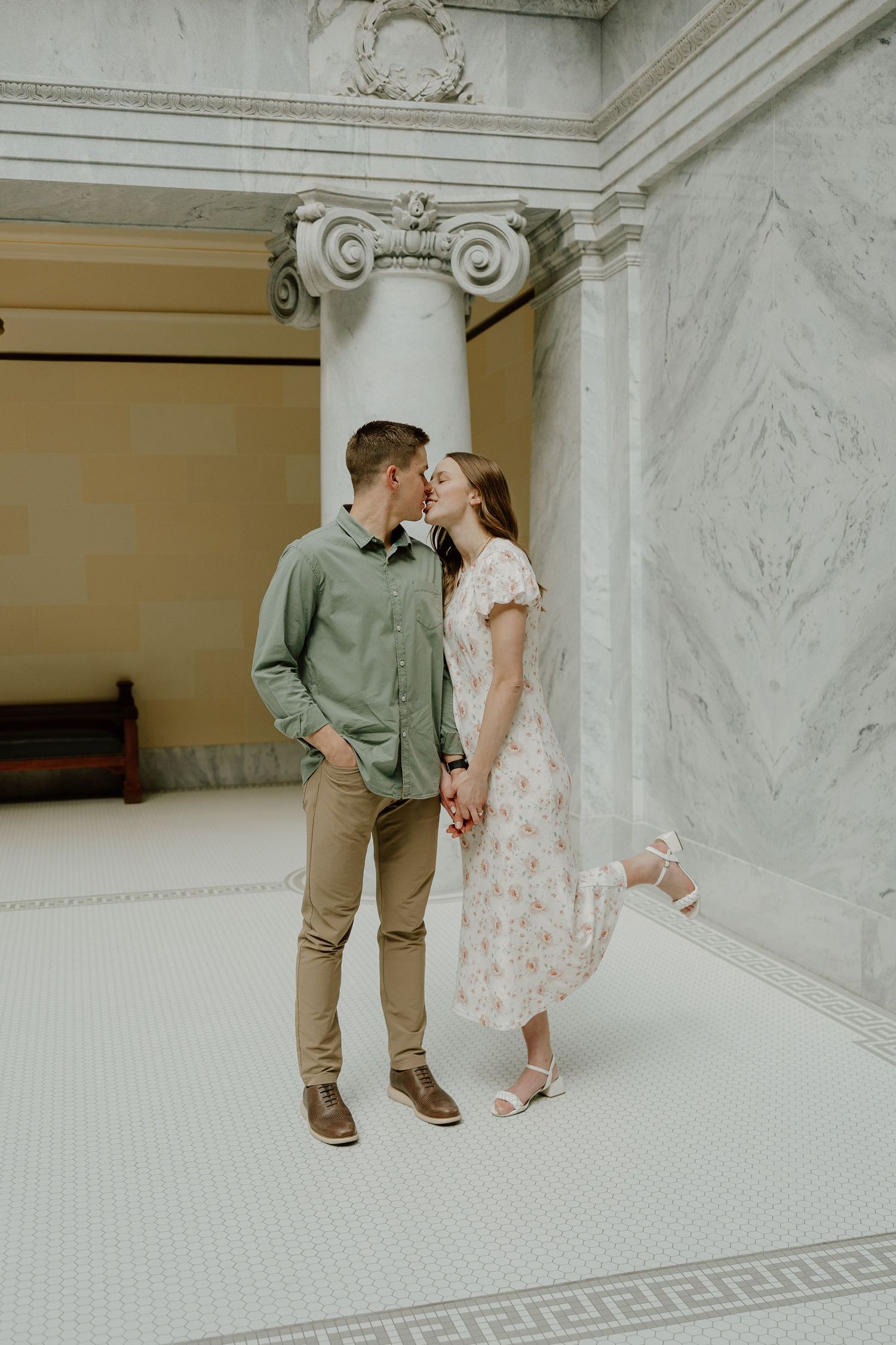 An engaged couple shares a kiss while holding hands beside ornate marble columns in a classical building.