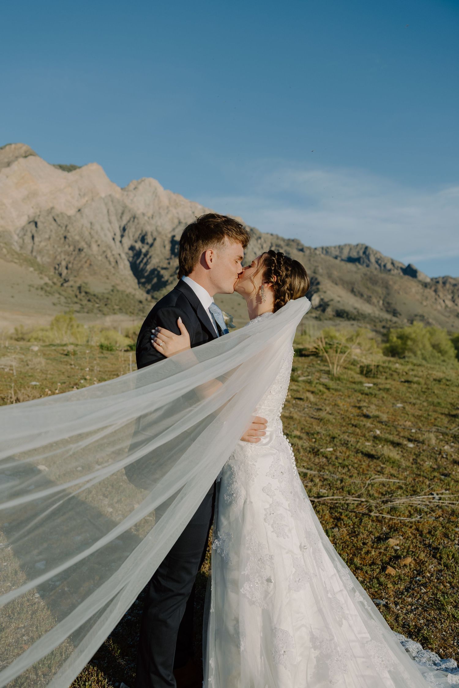 Bride and groom sharing a kiss with veil flowing in the wind against mountain scenery.