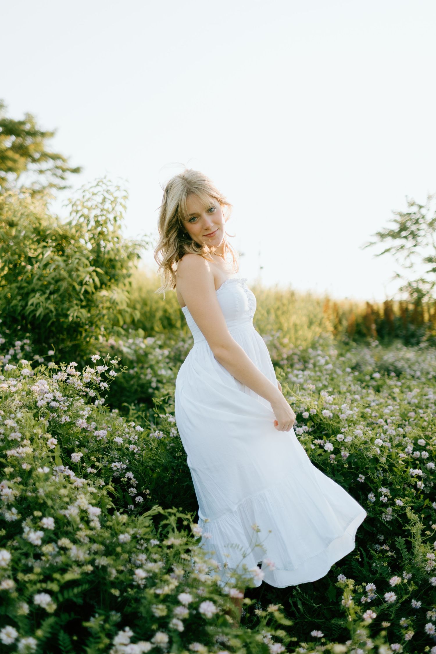 Young woman in white maxi dress standing in wildflower meadow in golden sunlight.