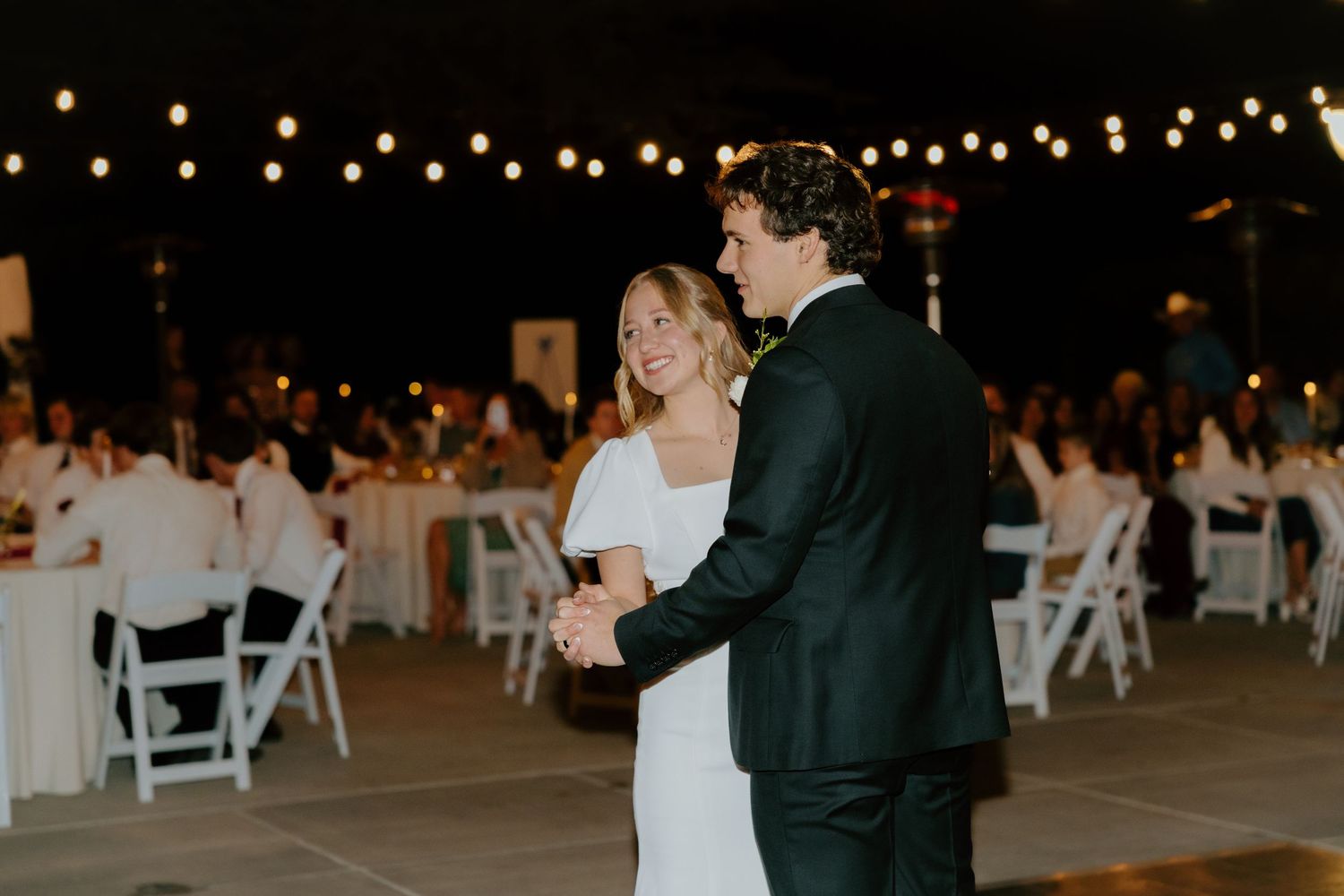Bride and groom share their first dance under string lights at an elegant outdoor wedding reception at night.