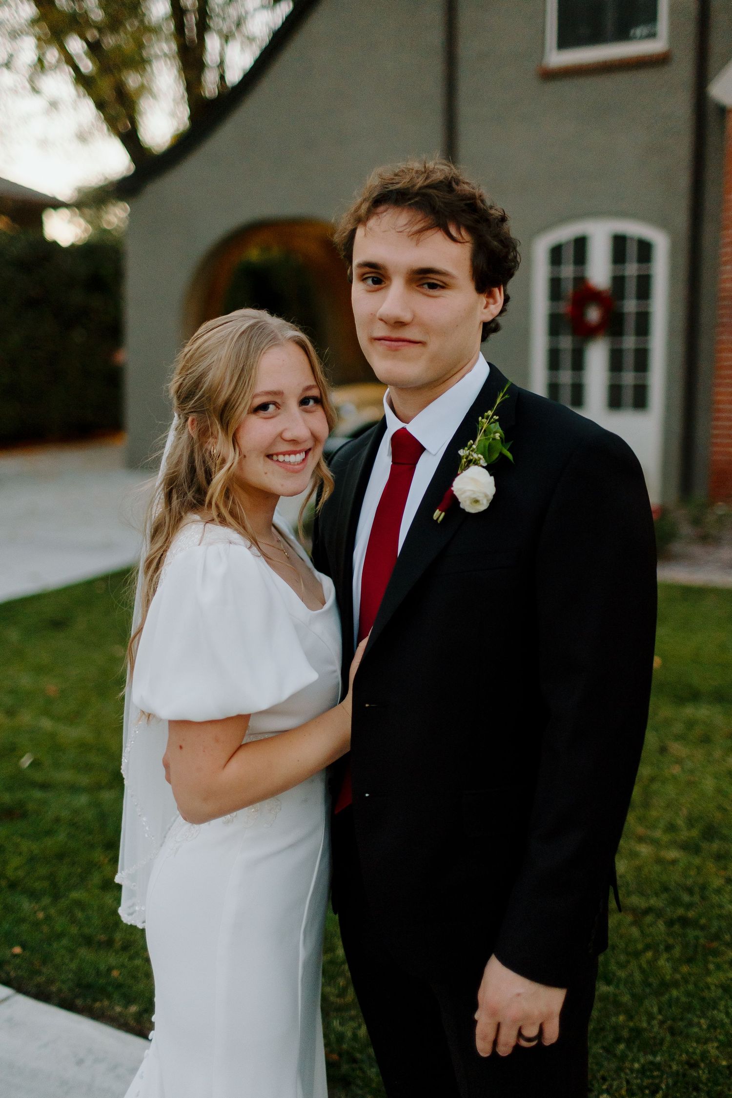 Bride in white dress and groom in black suit with red tie pose together outside a brick home.