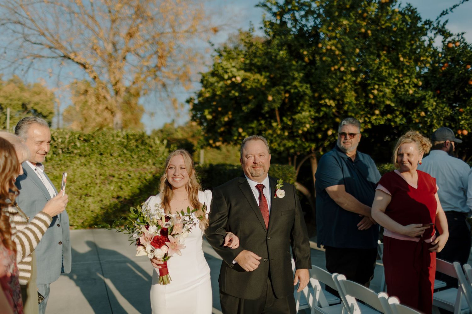 Bride walks down outdoor wedding aisle escorted by father in gray suit, guests seated on white chairs beside them.