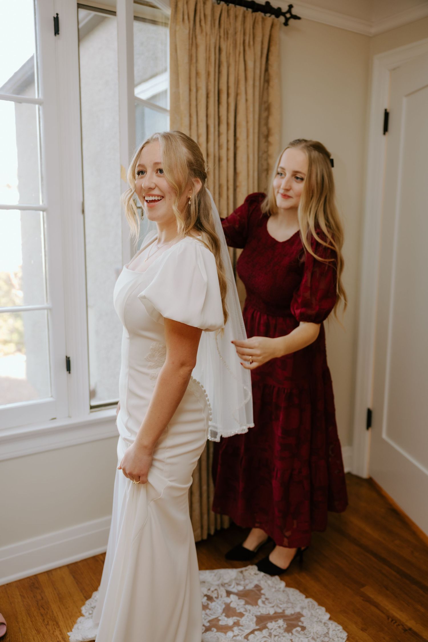 Bridesmaid in burgundy dress helps bride in white flutter-sleeve gown with her veil near a bright window.
