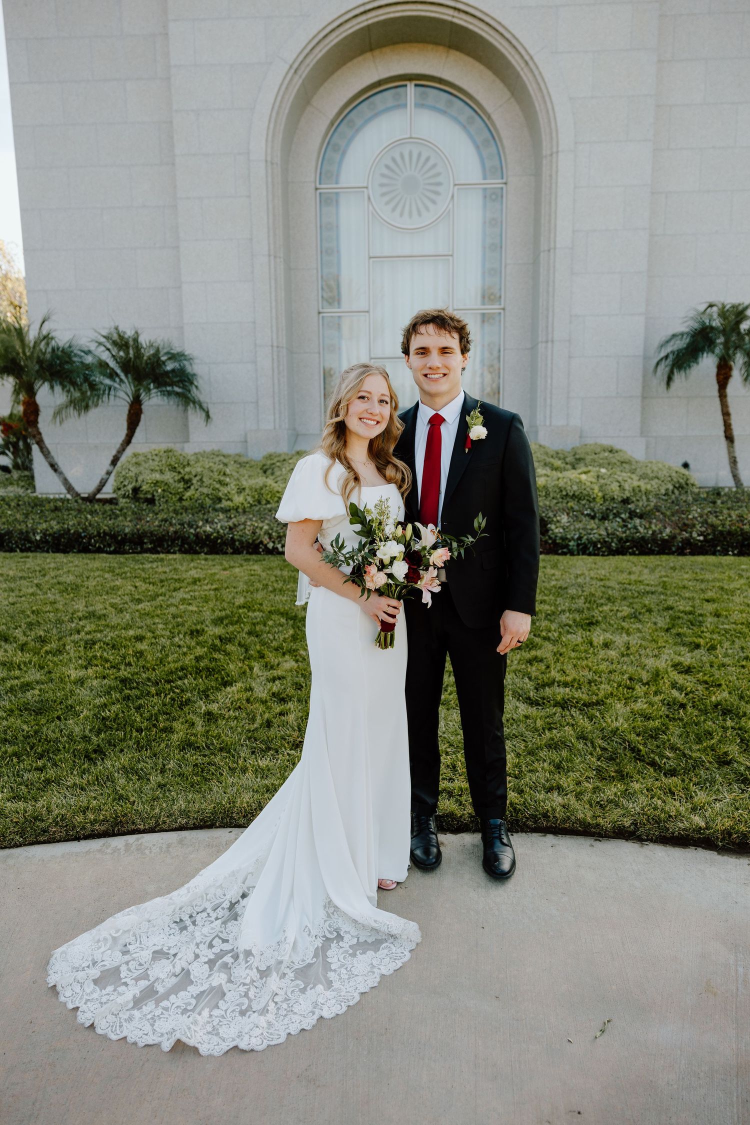 Bride in white lace gown and groom in black suit with red tie pose outside a temple on their wedding day.