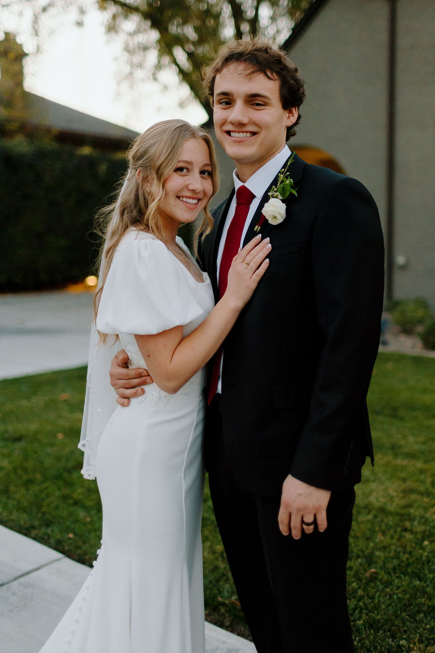 Smiling bride in white gown and groom in black suit with red tie pose together outdoors at their wedding.