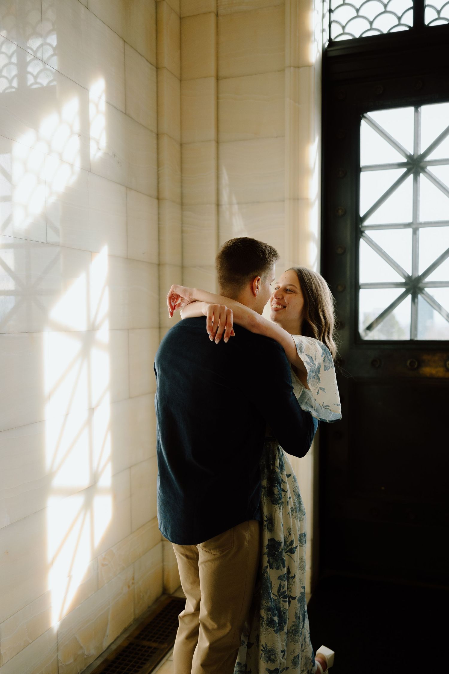 Couple embracing tenderly in soft sunlight near an ornate window, woman in floral dress smiling warmly at her partner.