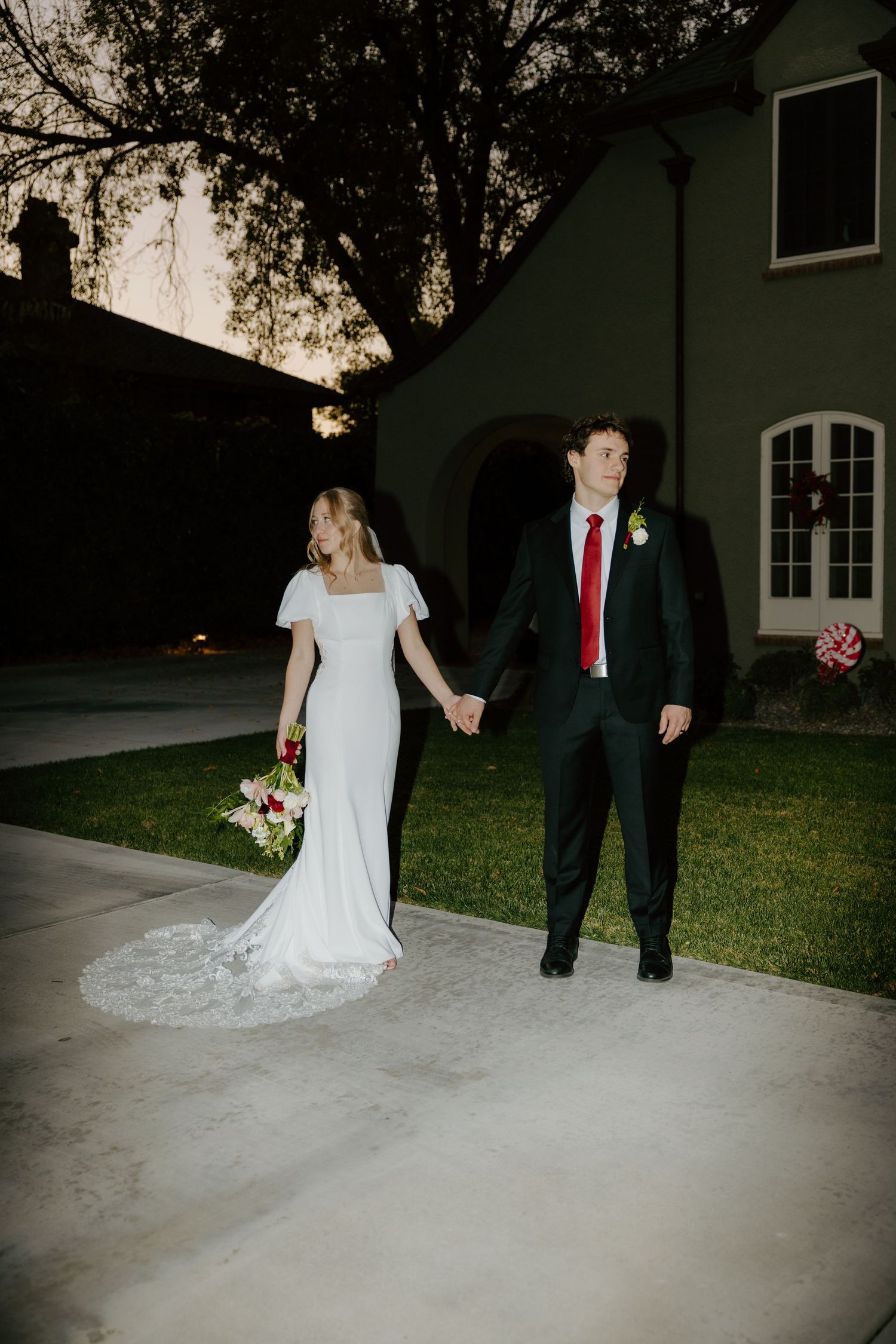 Bride and groom holding hands at dusk outside elegant venue, bride in white gown with bouquet, groom in dark suit with red tie.