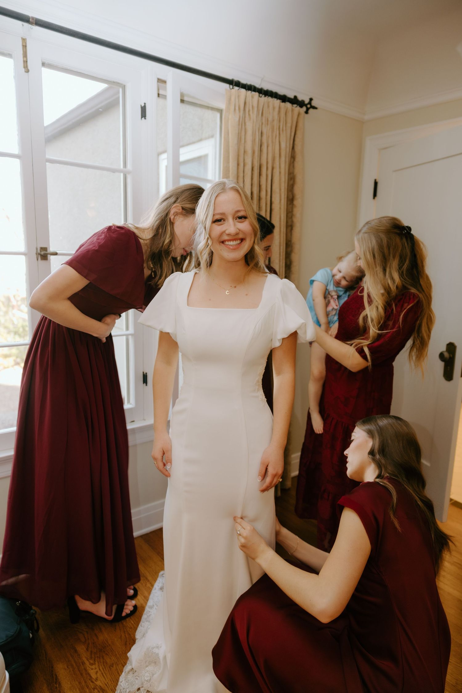 Smiling bride in white gown is surrounded by bridesmaids in burgundy dresses helping her get ready indoors.