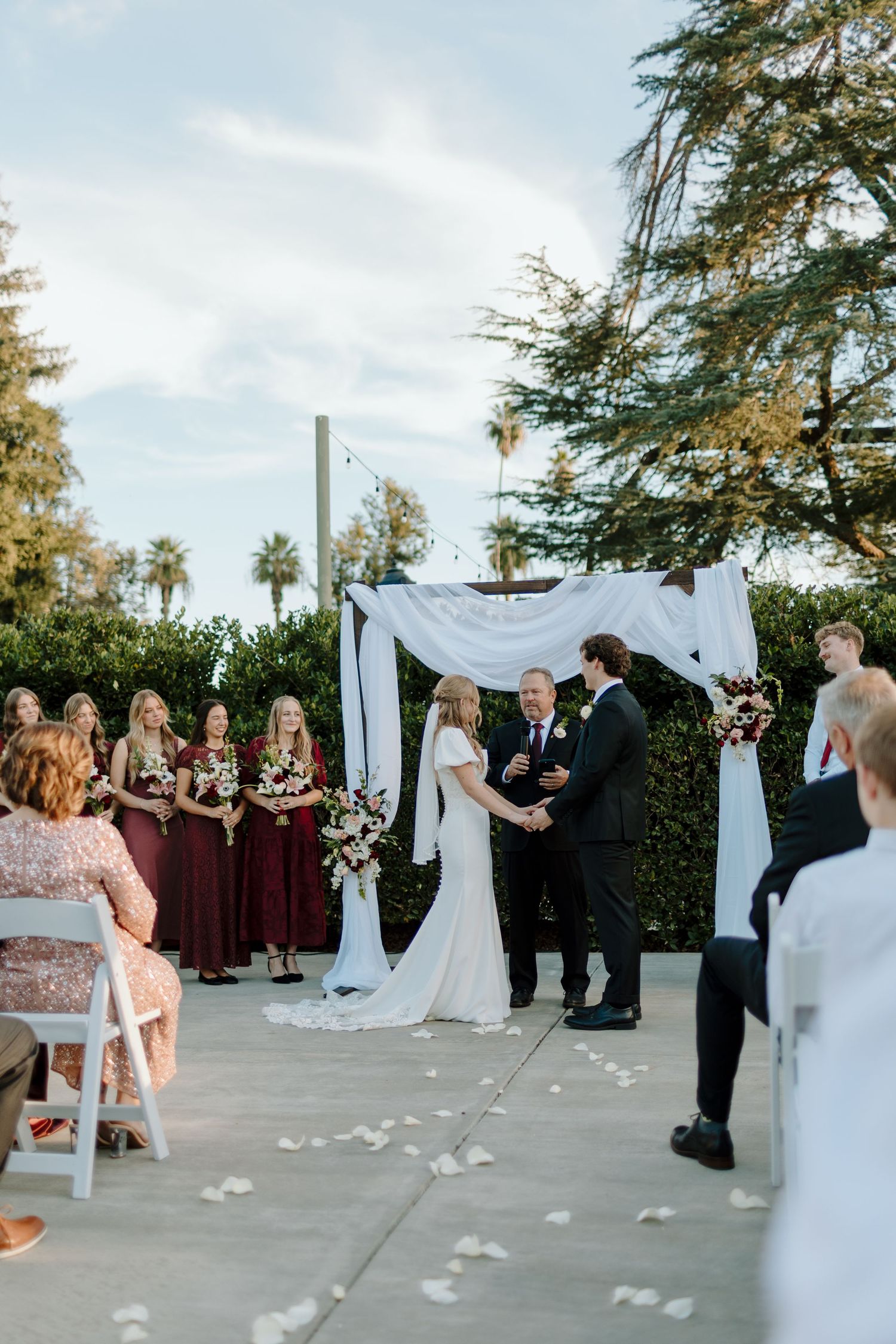 Outdoor wedding ceremony with bride and groom at floral arch, bridesmaids in burgundy dresses, white rose petal aisle.