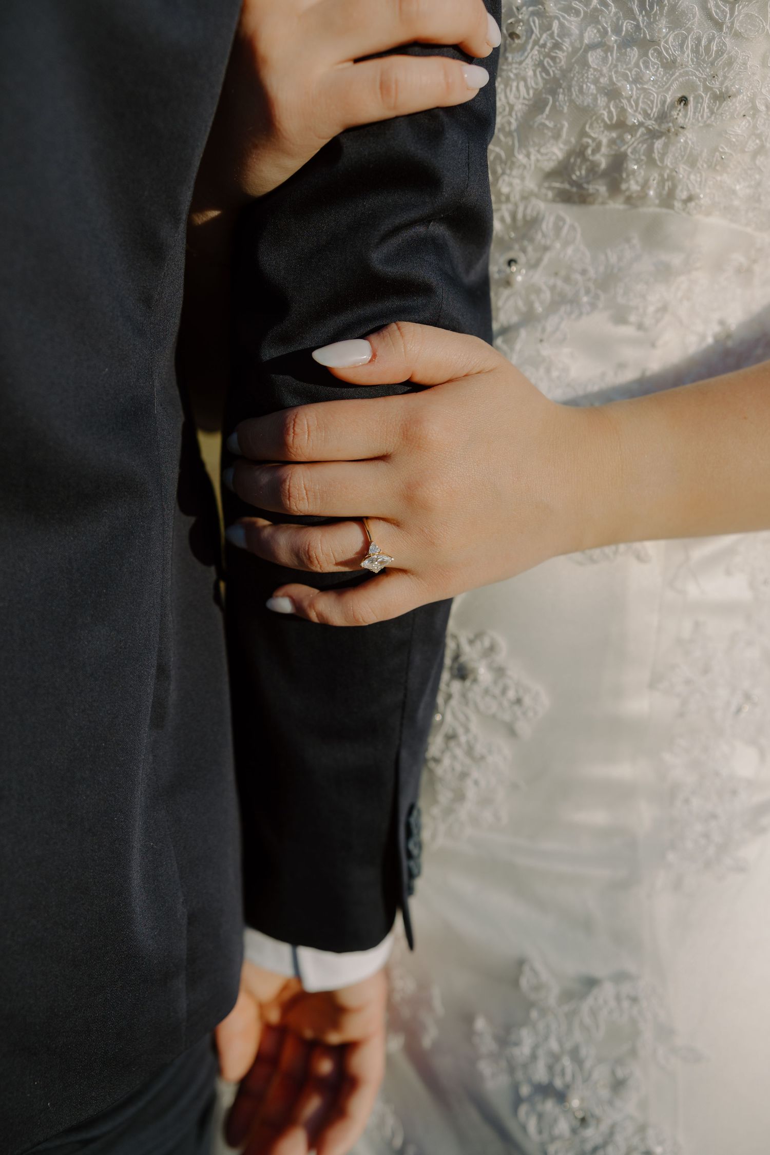 Close-up of bride's hand with diamond engagement ring resting on groom's black suit sleeve.