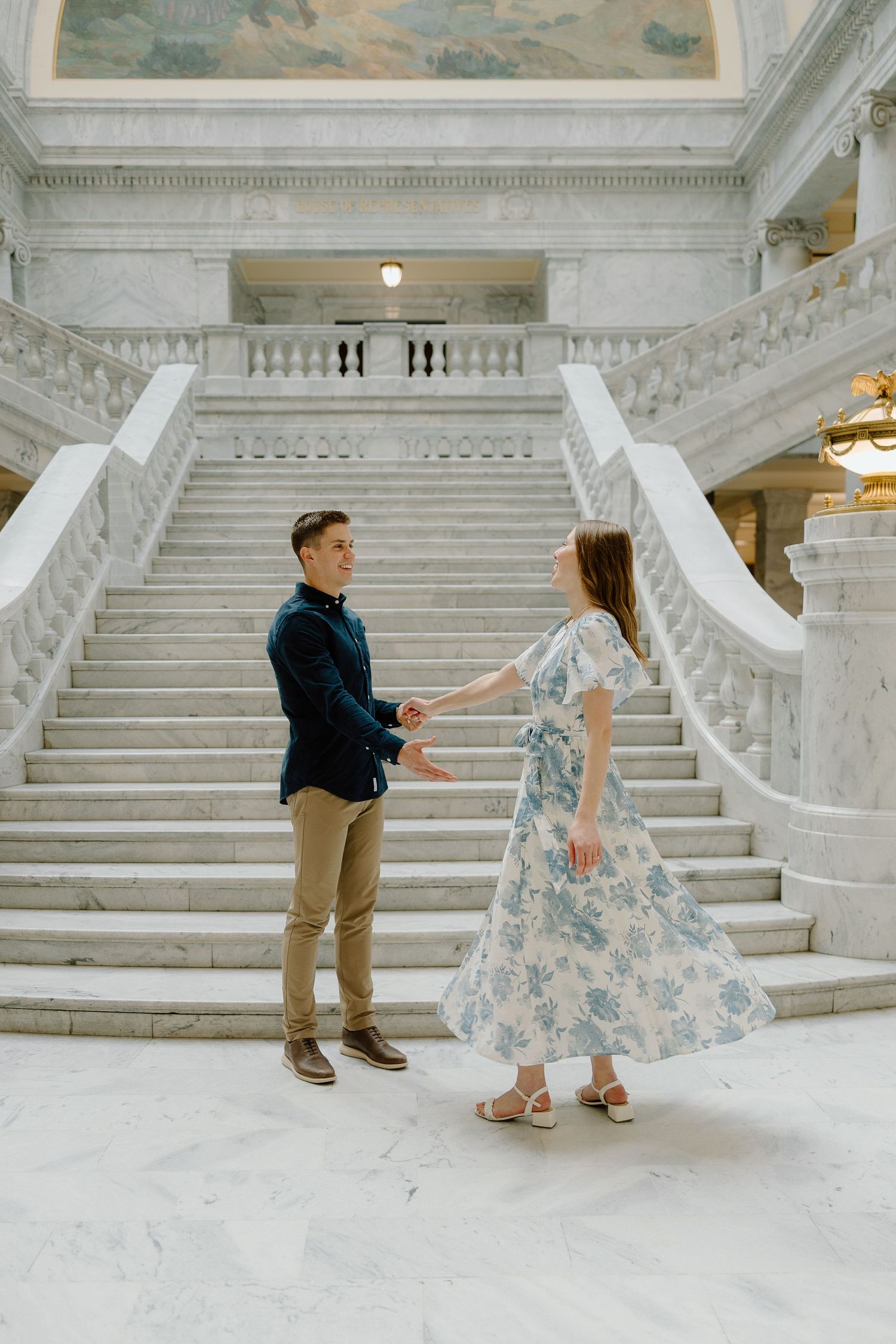 Couple holding hands and dancing at the base of grand marble staircase in elegant classical building.