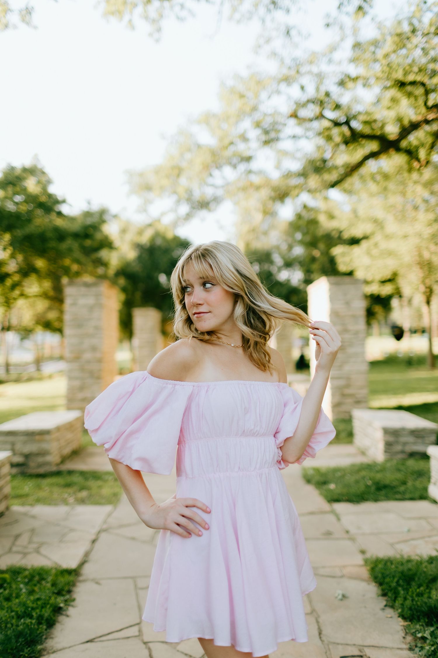 Young woman in pink off-shoulder puff sleeve mini dress posing outdoors in a park with stone pillars.