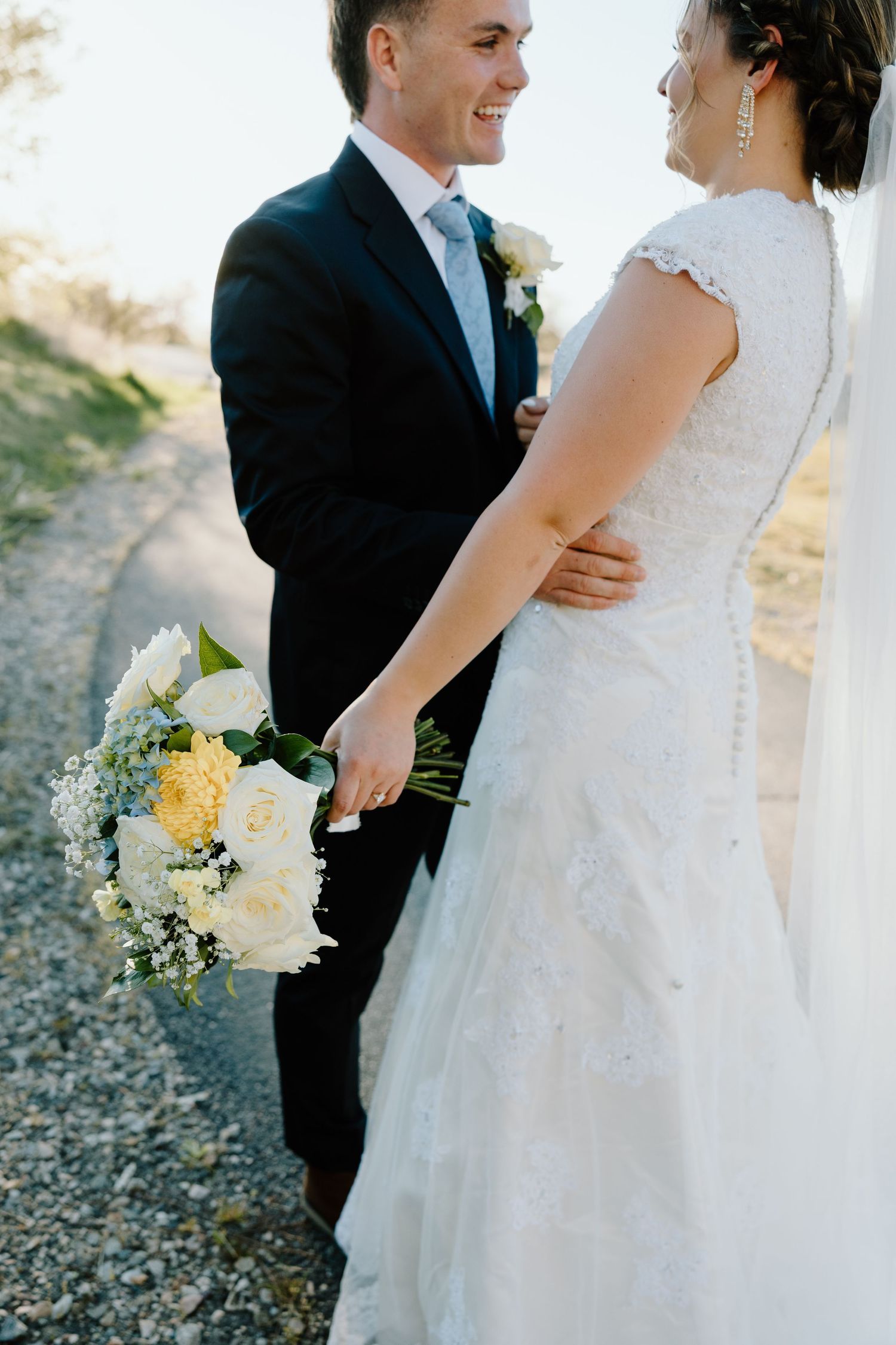 Smiling groom in black suit holds white rose bouquet while embracing bride in lace wedding dress outdoors.