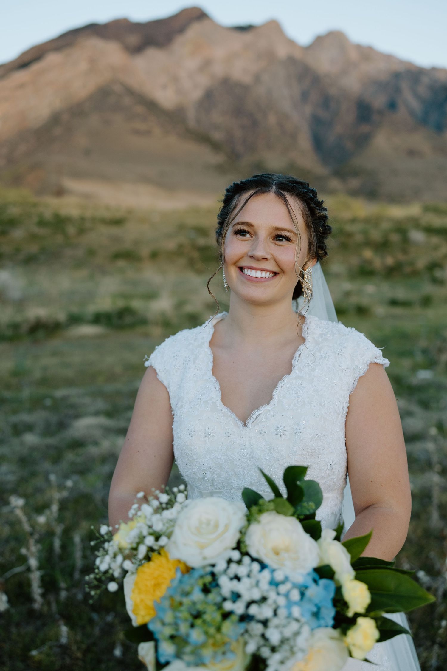 Smiling bride in lace wedding dress holds blue and yellow bouquet with stunning mountain scenery behind her.