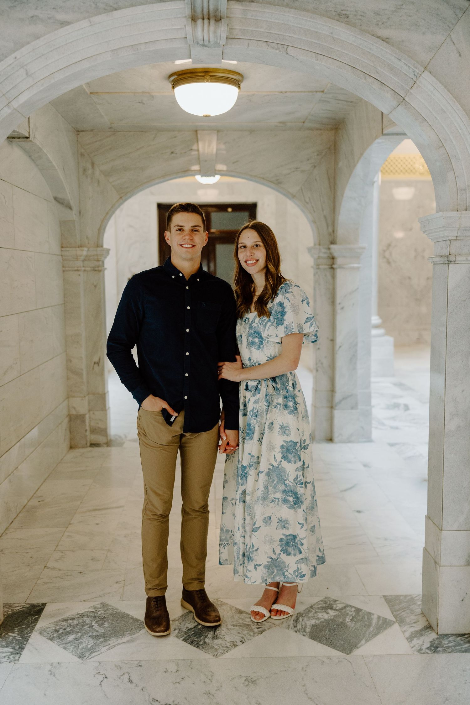 A smiling couple poses in an elegant marble archway corridor, man in navy shirt and khakis, woman in floral dress.