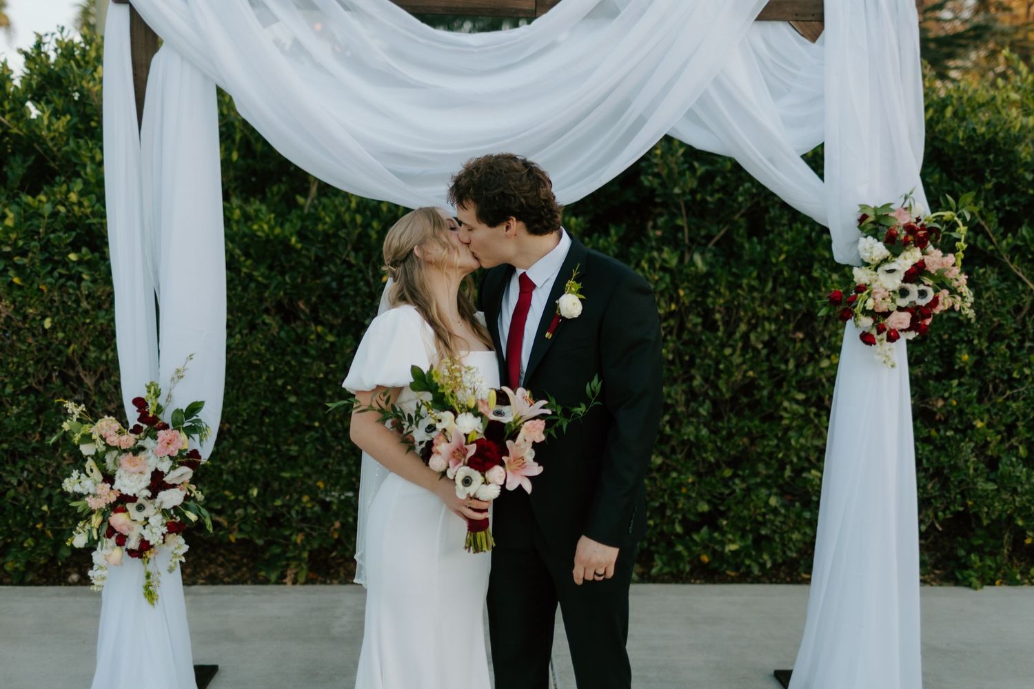 Bride and groom share first kiss under white draped floral arch decorated with pink and red flowers.