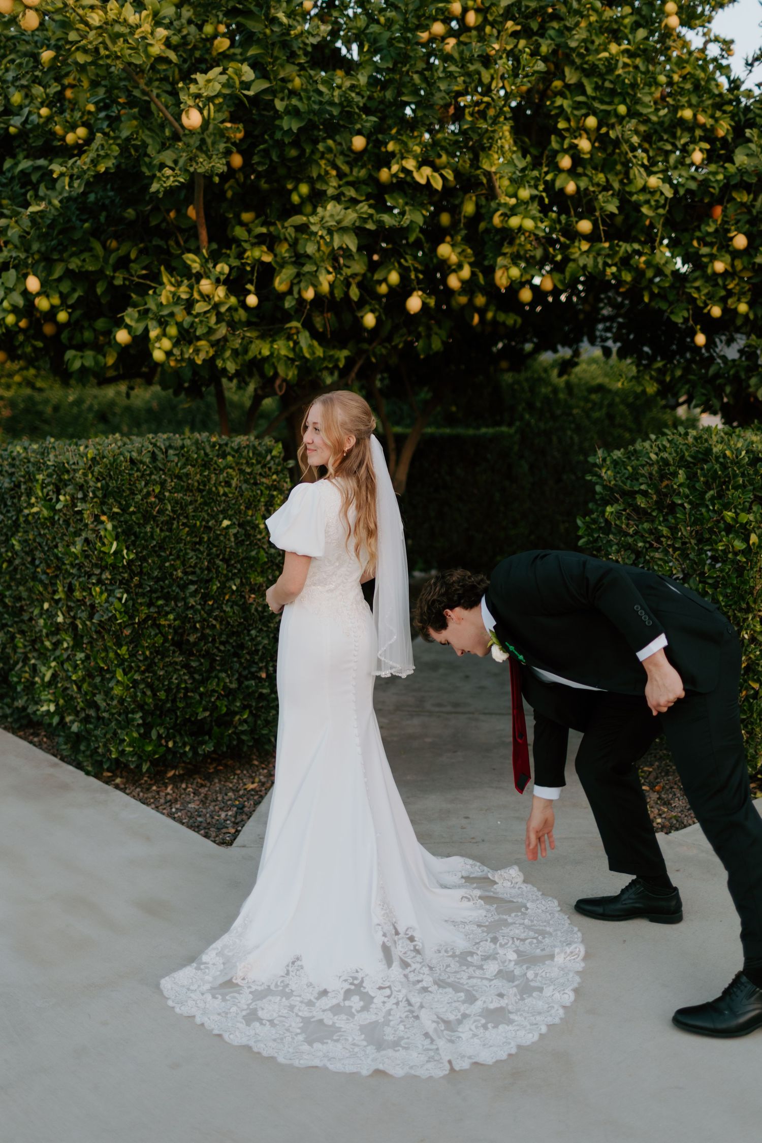 Bride in elegant lace-train white gown stands near lemon tree as groom adjusts her wedding dress train.