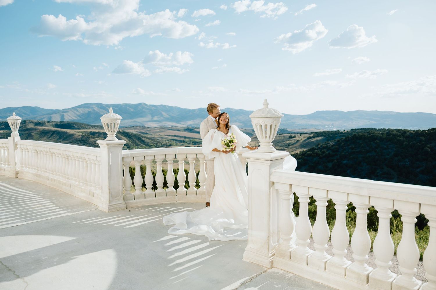 Bride and groom embrace on elegant white balcony with stunning mountain landscape backdrop on sunny wedding day.