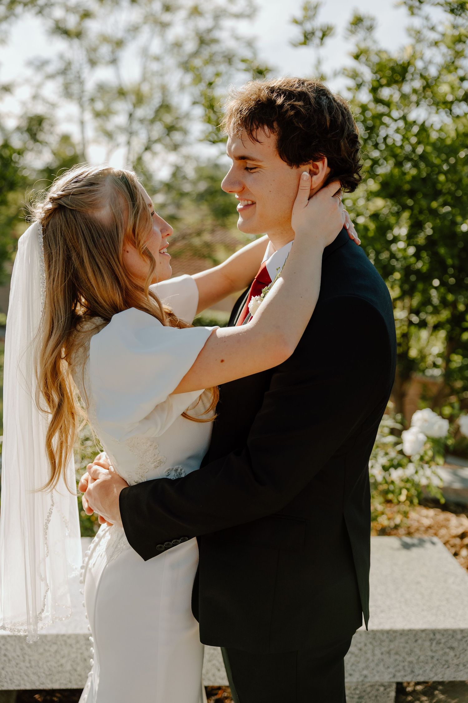 A bride and groom embrace outdoors, smiling at each other, she in a white gown with veil, he in a dark suit.