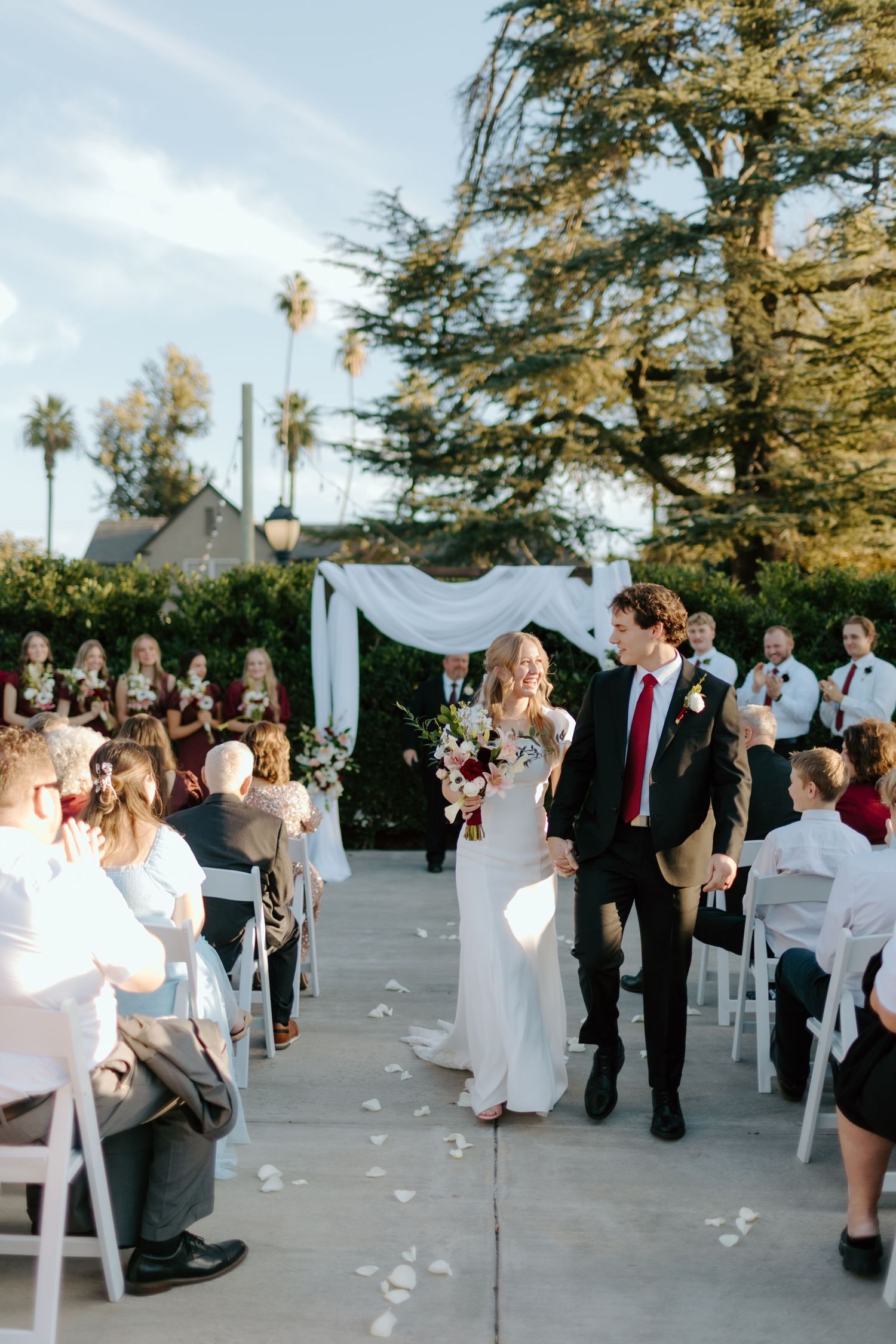 Bride and groom walk down outdoor wedding aisle, guests seated in white chairs, floral arch and tall trees in background.