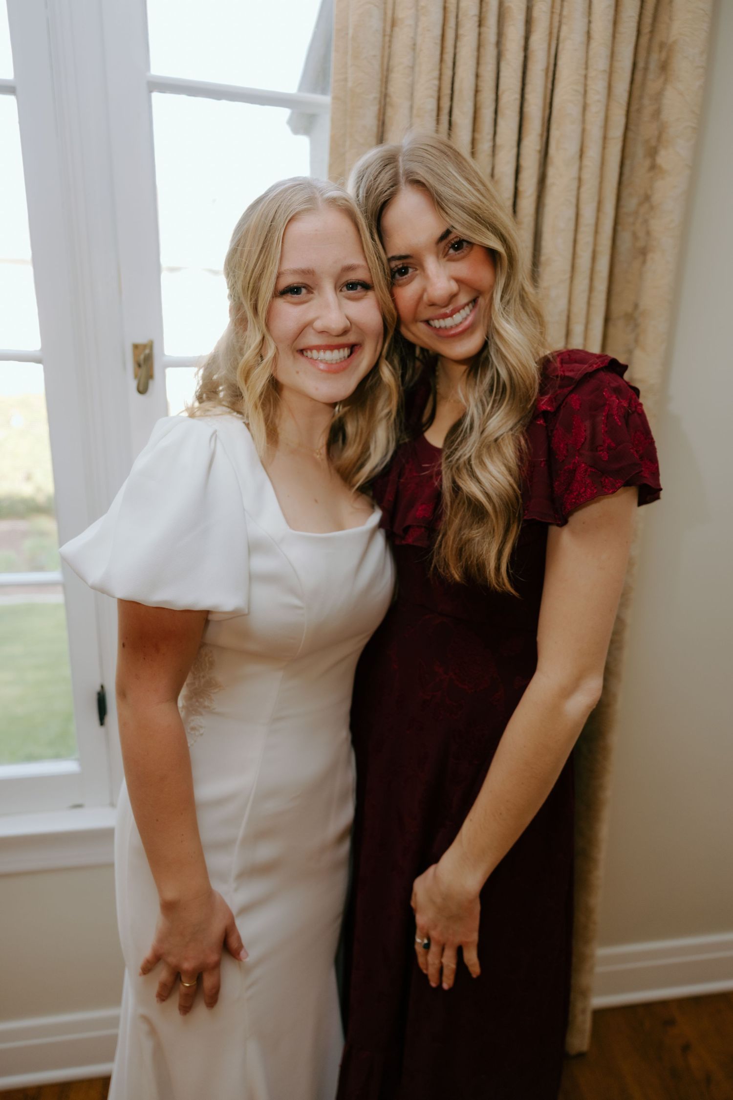 Two blonde women smile together, one in a white wedding dress and one in a burgundy floral dress.