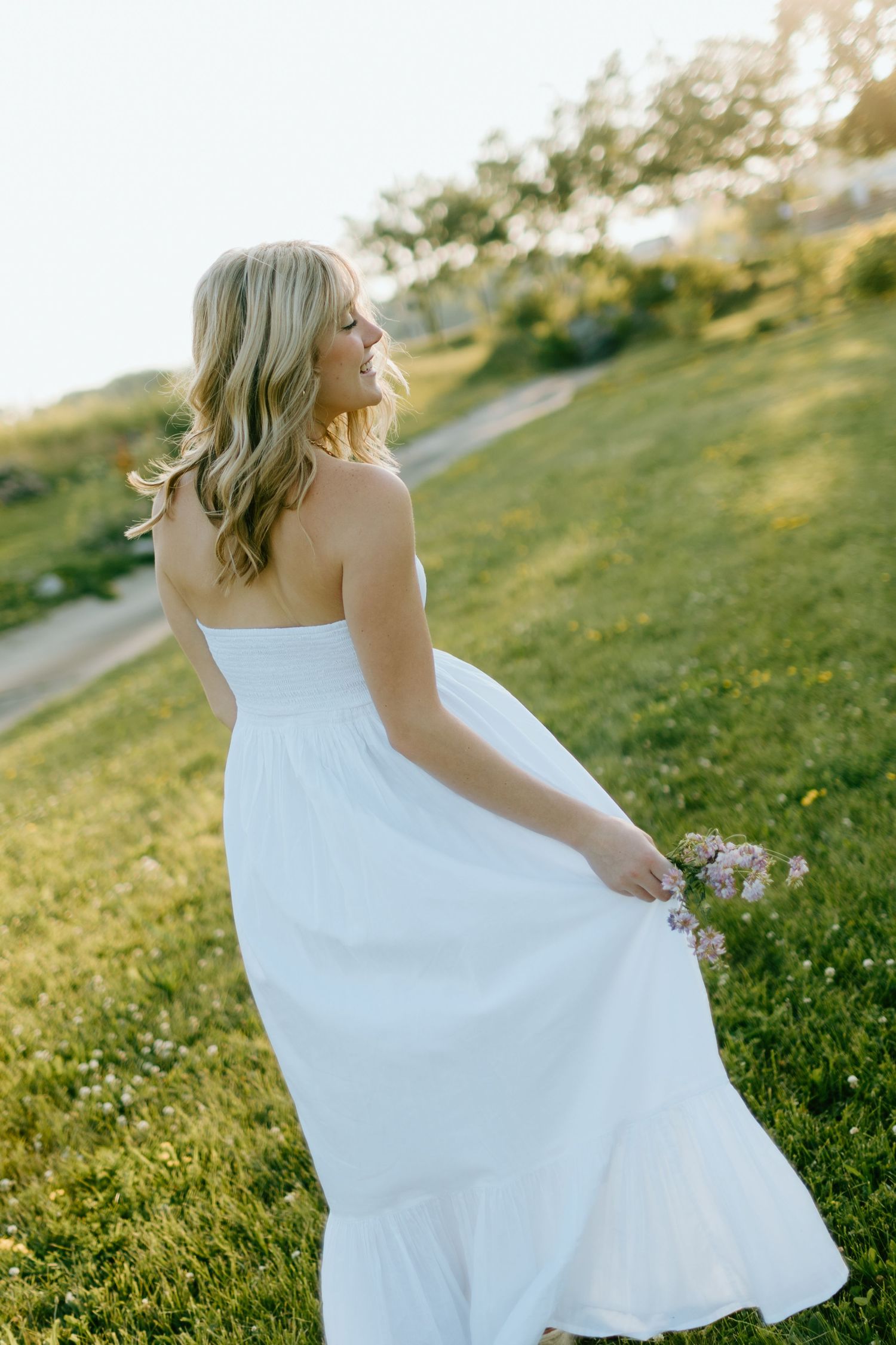 Blonde woman in white strapless dress holding flowers, standing on green grass in golden sunlight.