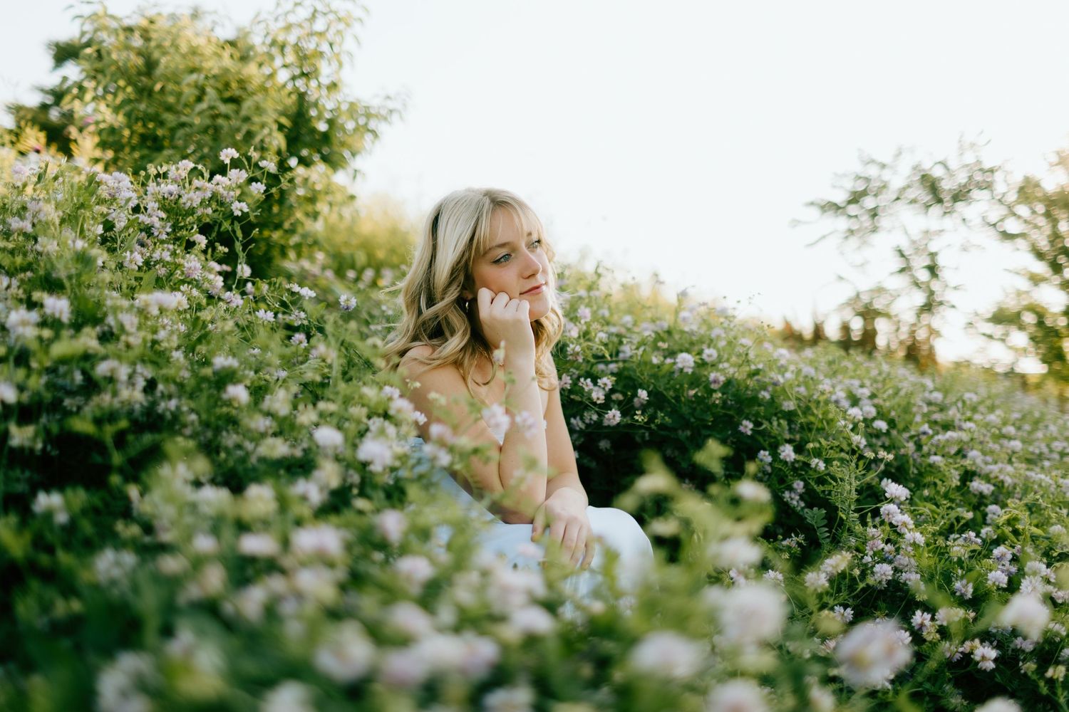 Woman sitting among blooming white wildflowers in a sunlit garden, wearing a floral dress with a serene expression.