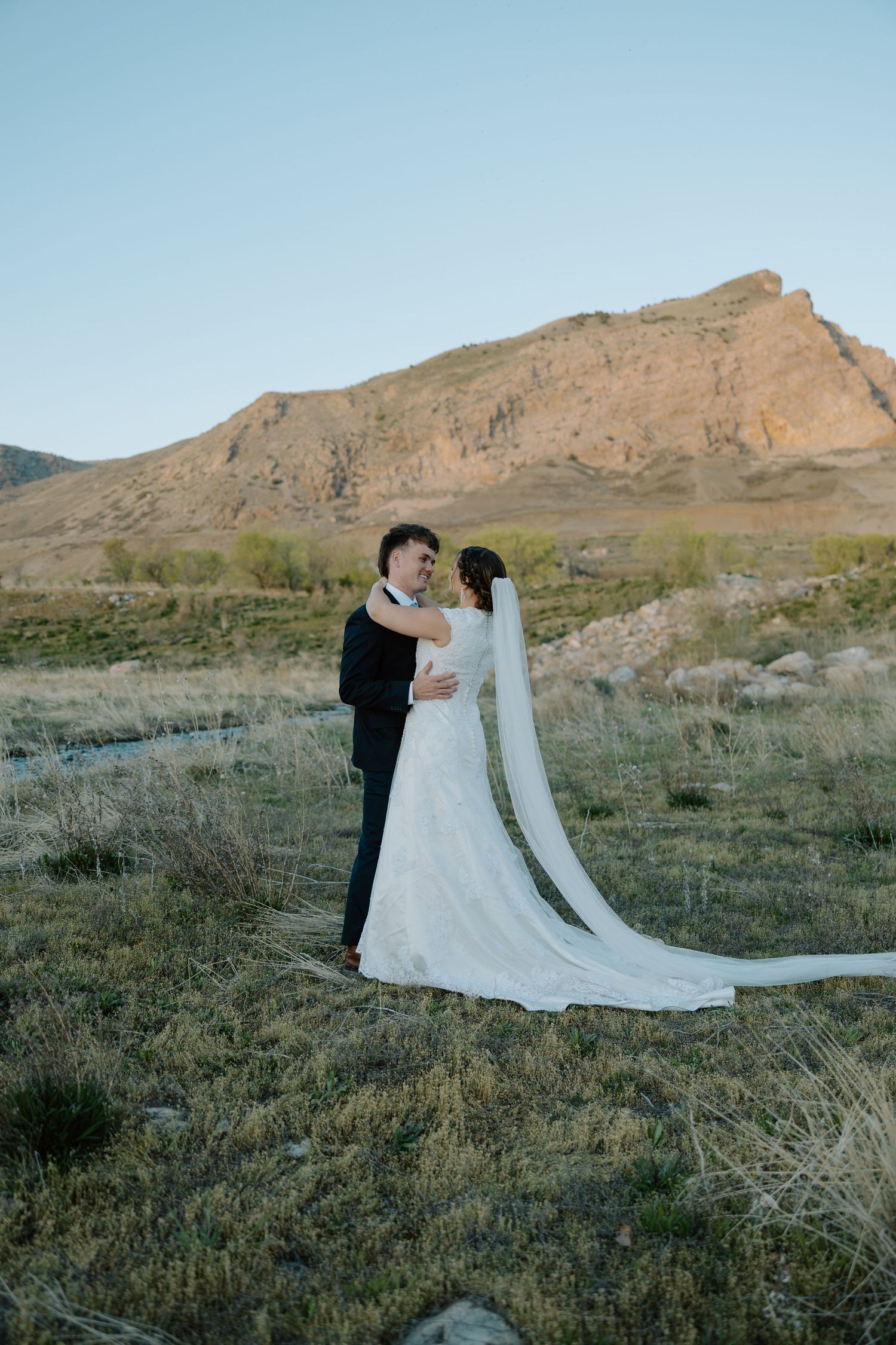 Bride and groom embrace in a scenic outdoor wedding portrait with golden mountain backdrop at dusk.
