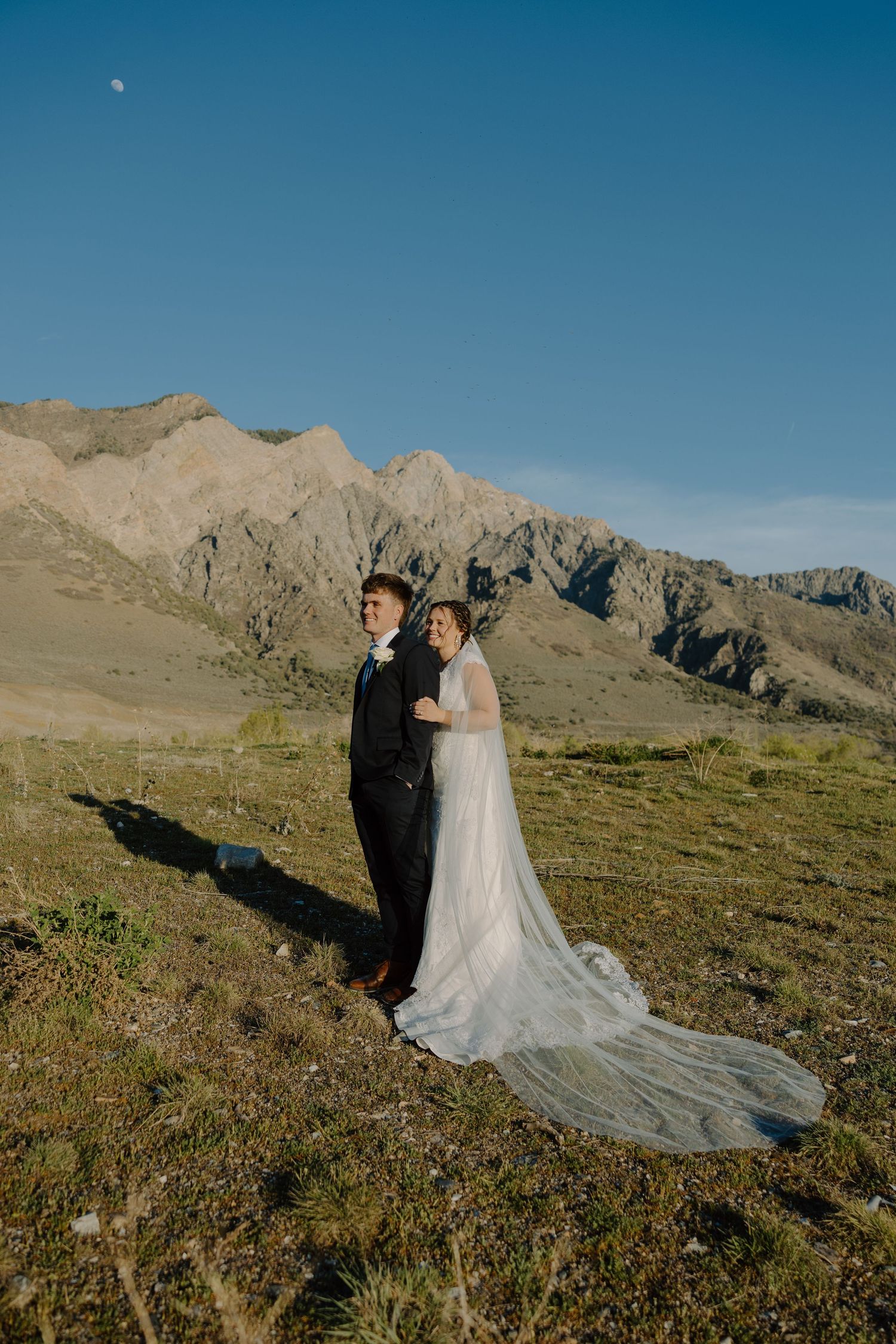 Bride and groom standing together in a scenic mountain landscape with a long flowing wedding veil.