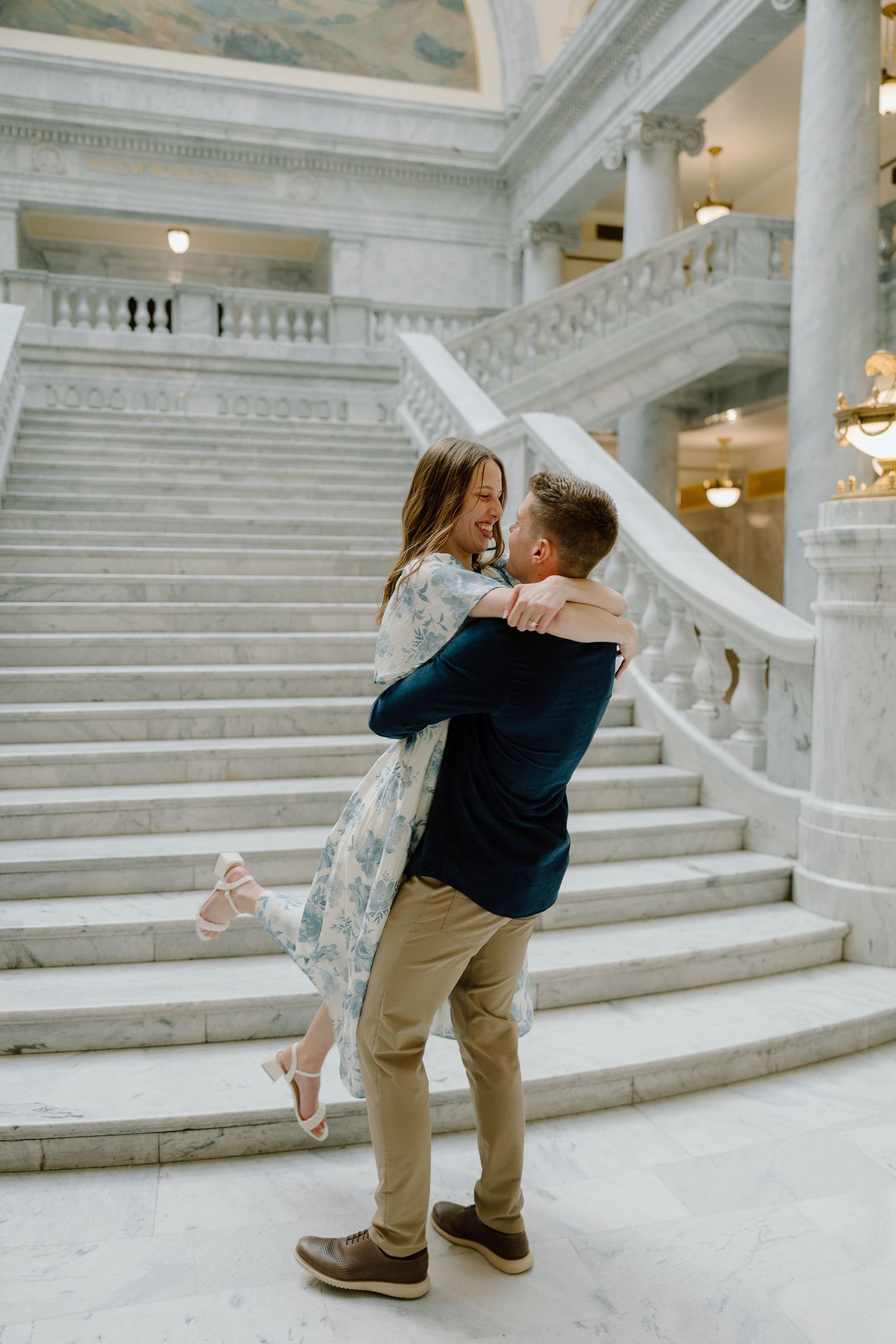 Couple embracing on marble stairs inside a grand capitol building, man lifting woman in floral dress.