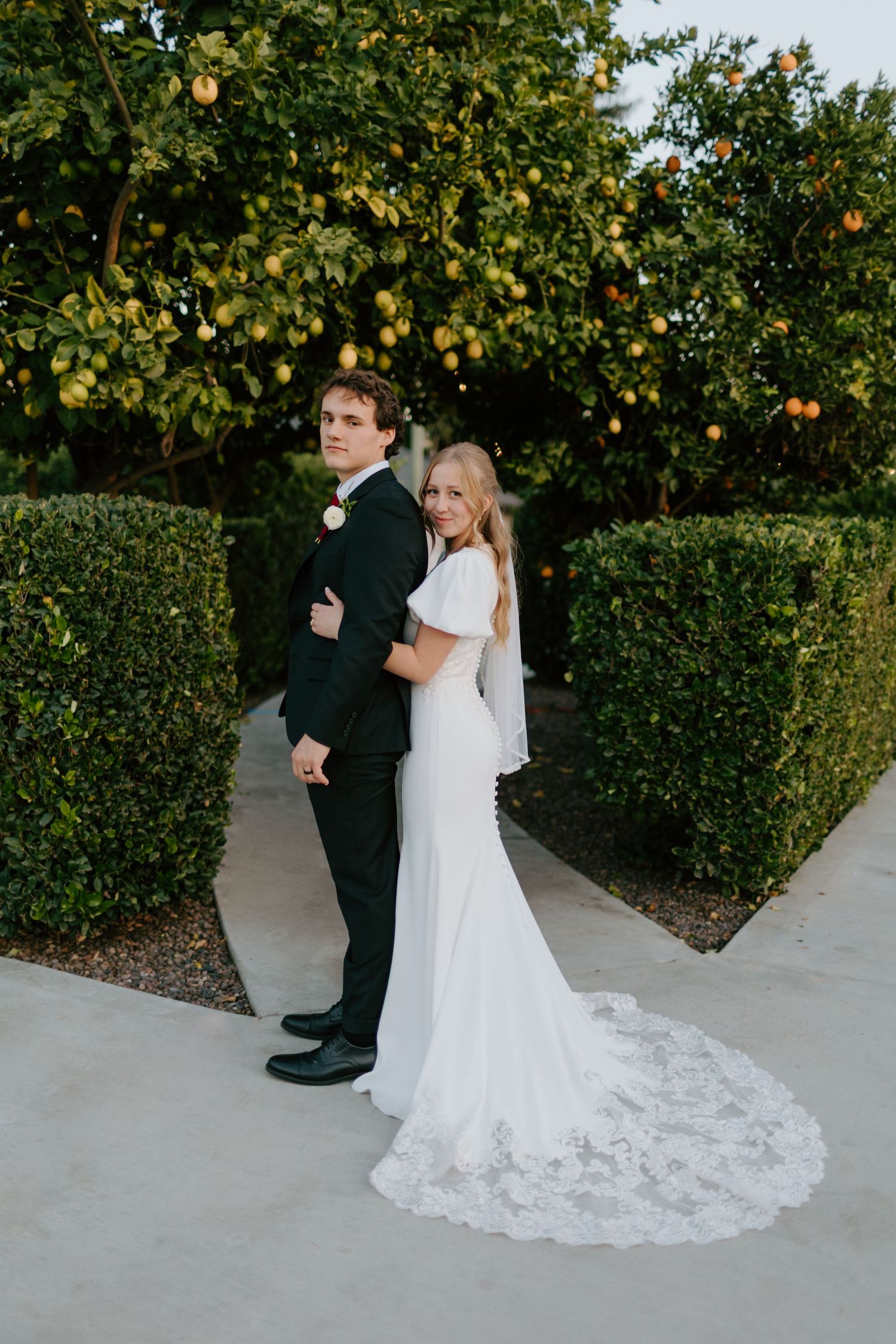 Bride and groom standing back-to-back in a lush garden with citrus trees, bride wearing a lace train wedding gown.