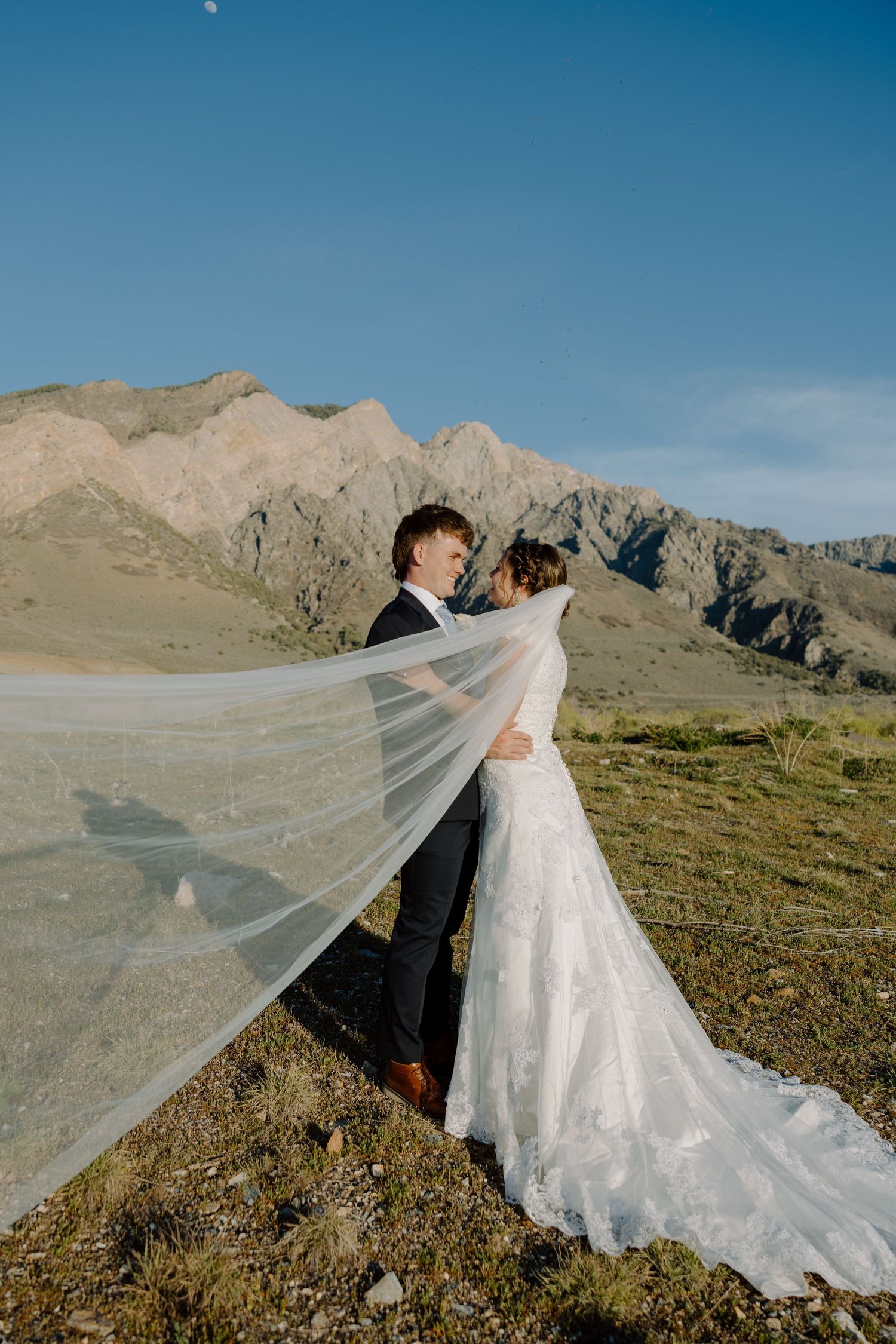 Bride's veil billows in the wind as couple embraces with stunning mountain scenery behind them.