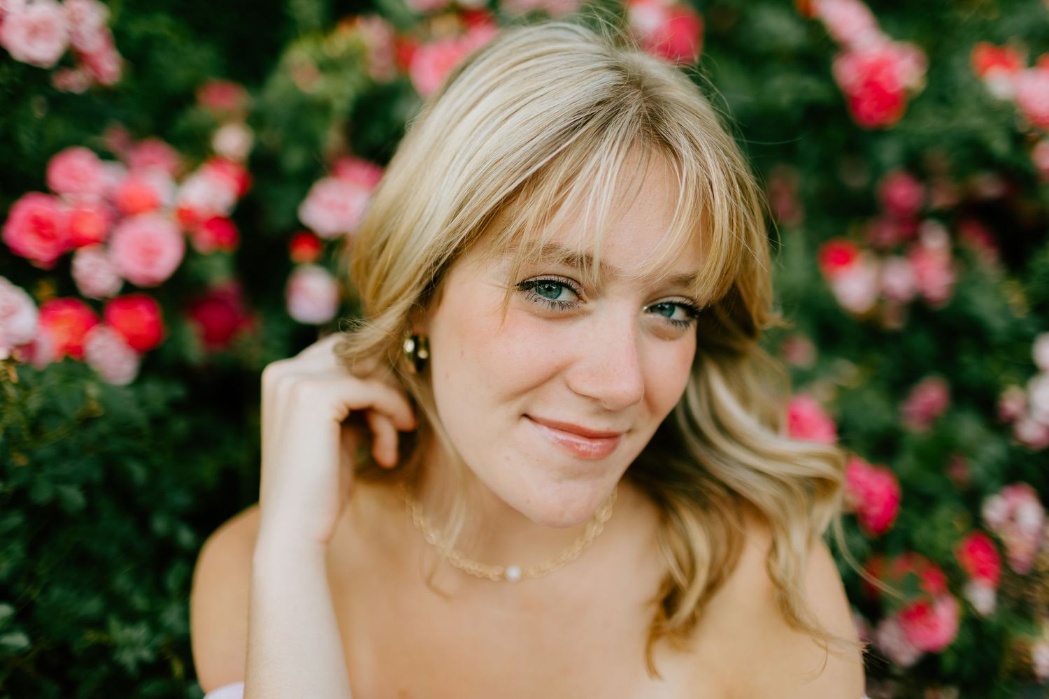 Smiling blonde woman with green eyes wearing a necklace, posing outdoors in front of blooming pink roses.
