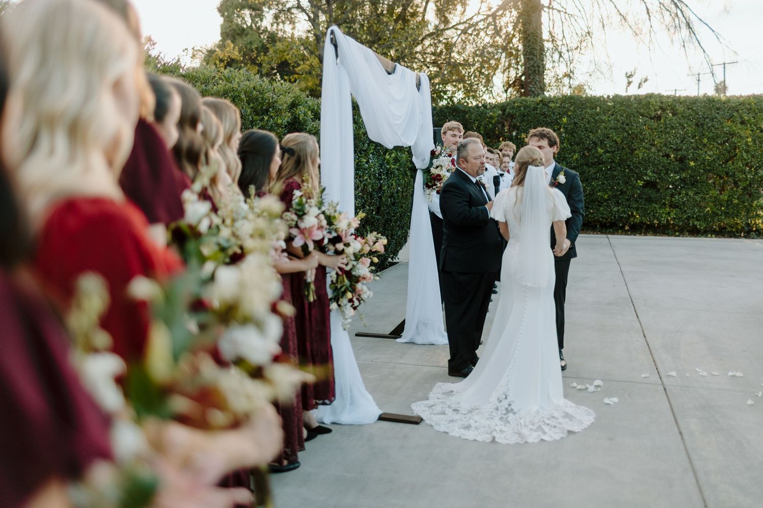 Outdoor wedding ceremony with bride and groom at floral arch, bridesmaids in burgundy dresses holding bouquets.