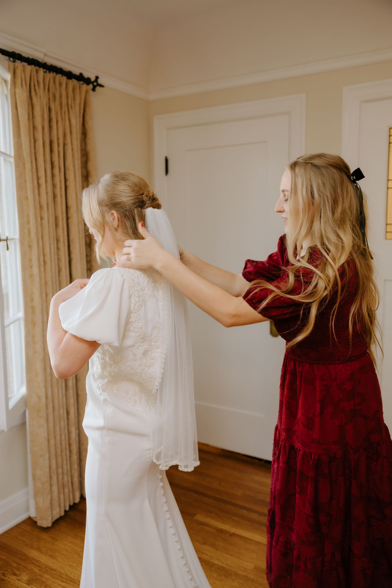 Bridesmaid in burgundy velvet dress adjusts veil on bride in white lace-back wedding gown indoors.