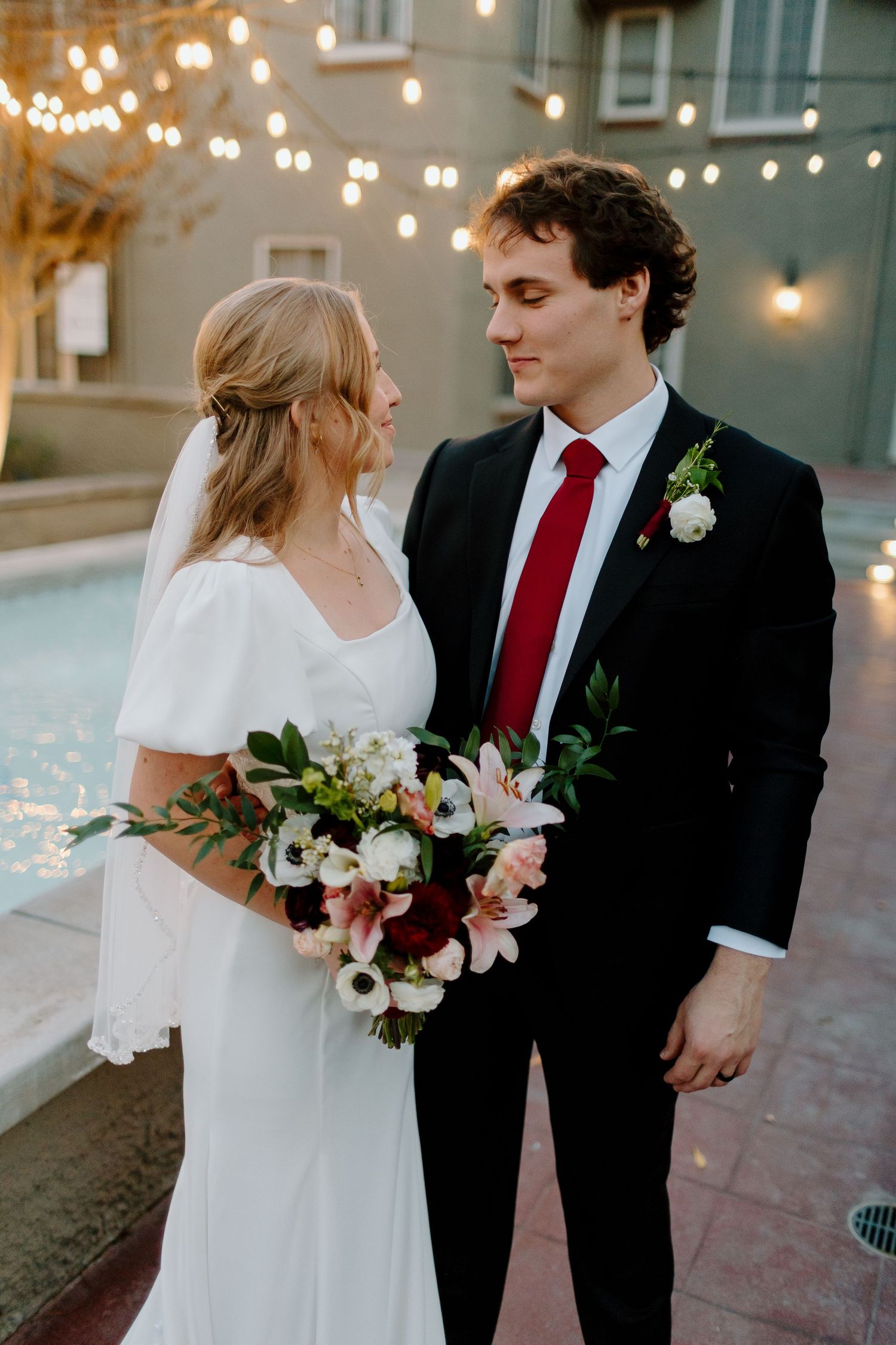 Bride and groom share a romantic moment outdoors, with string lights glowing behind them near a pool.