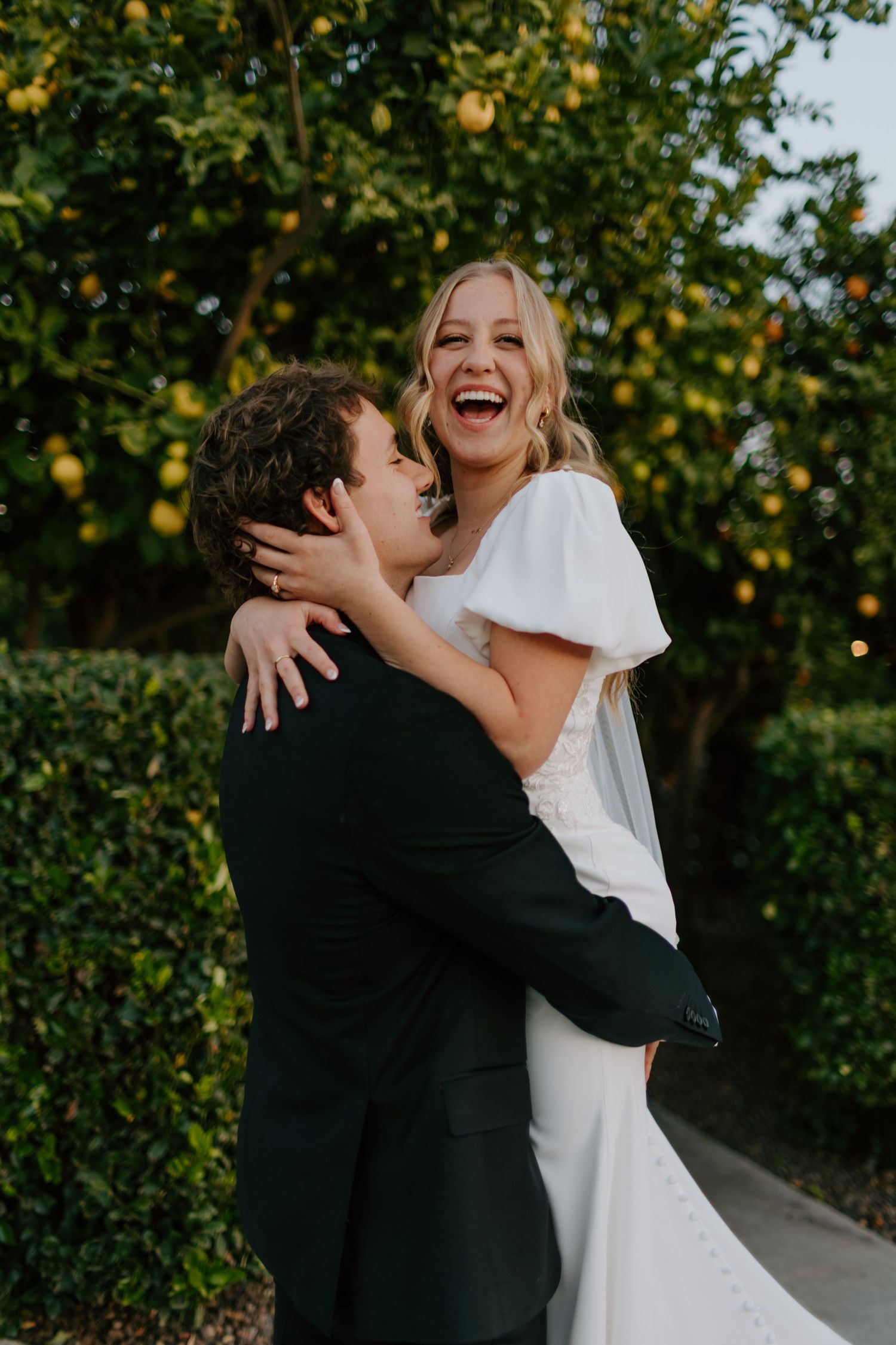 Joyful bride laughs while groom lifts her in a loving embrace beneath a lemon tree outdoors.