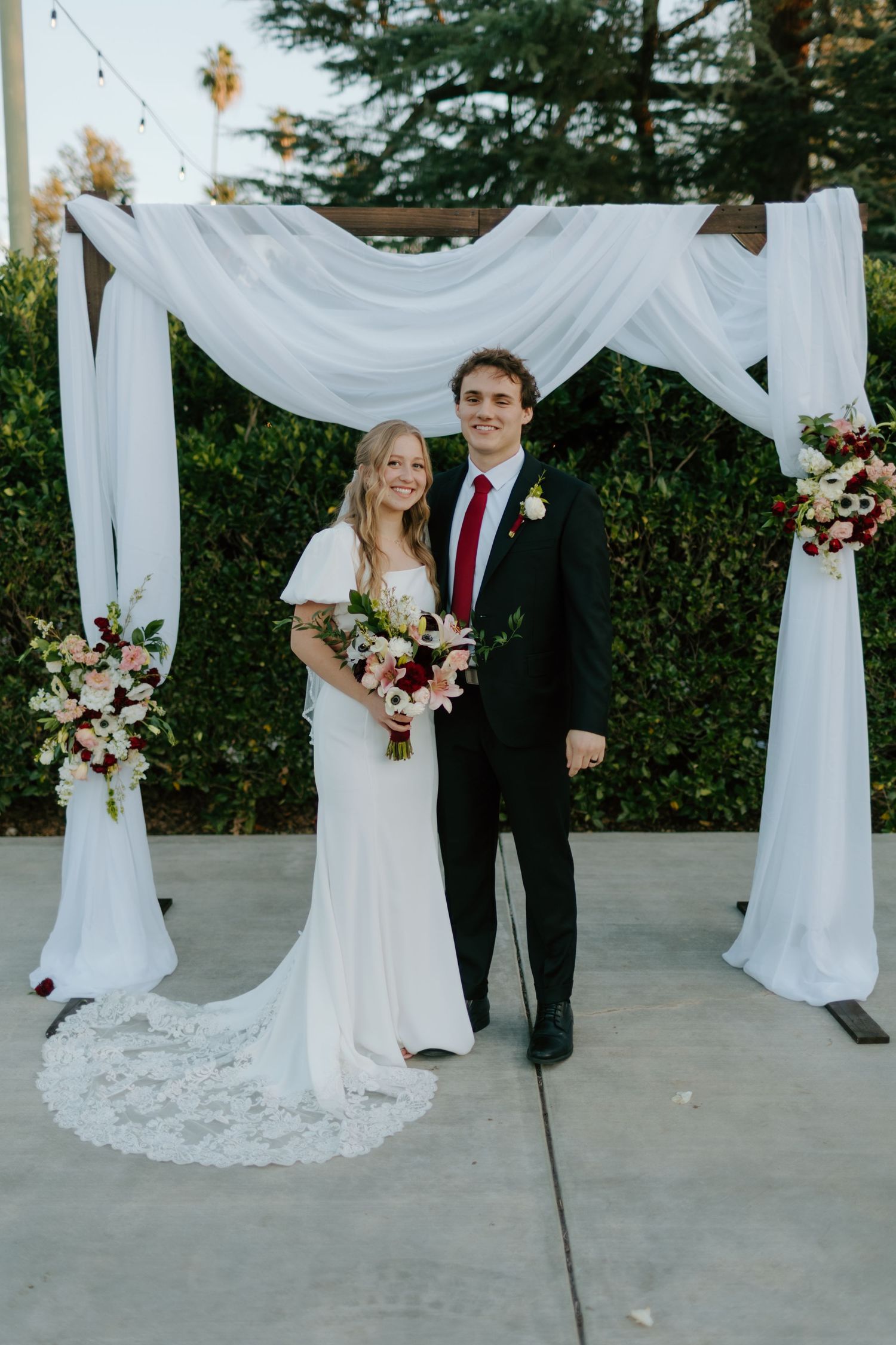Smiling bride and groom pose under a white draped floral arch with burgundy and blush flower arrangements.