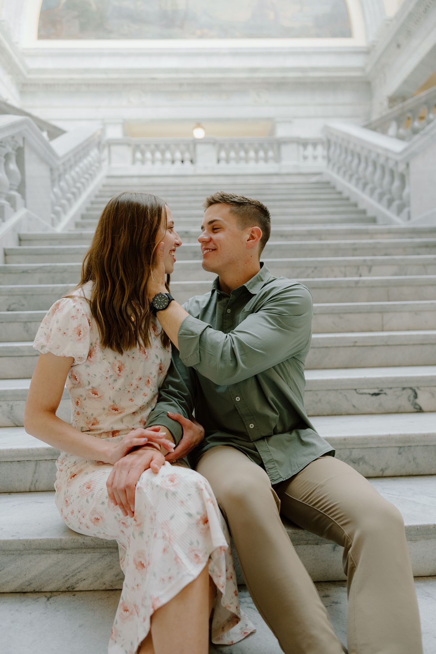 A couple sits on marble stairs gazing at each other, woman in floral dress and man in green shirt holding hands.
