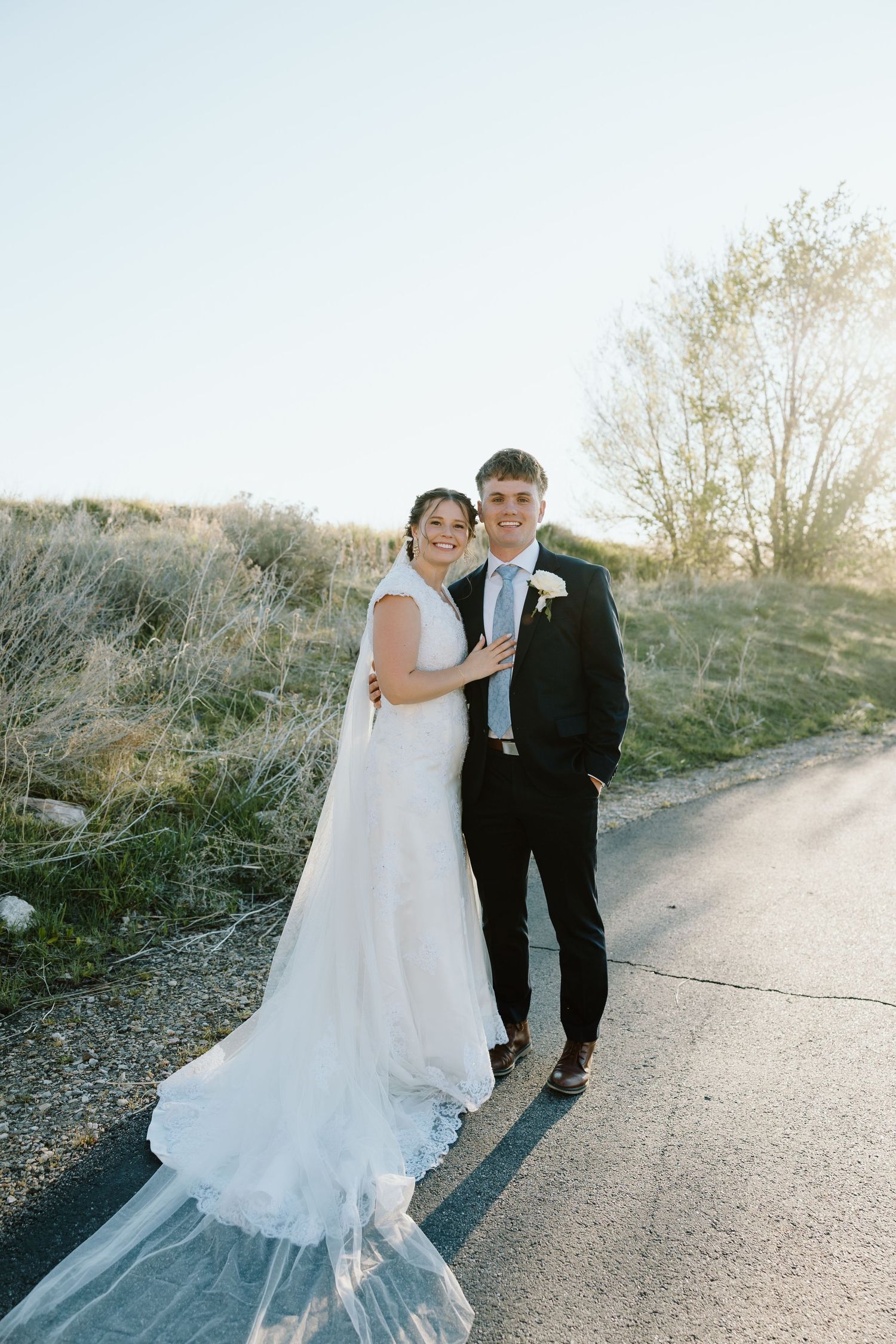 Bride in white gown with veil and groom in black suit pose together on a sunlit road with greenery.