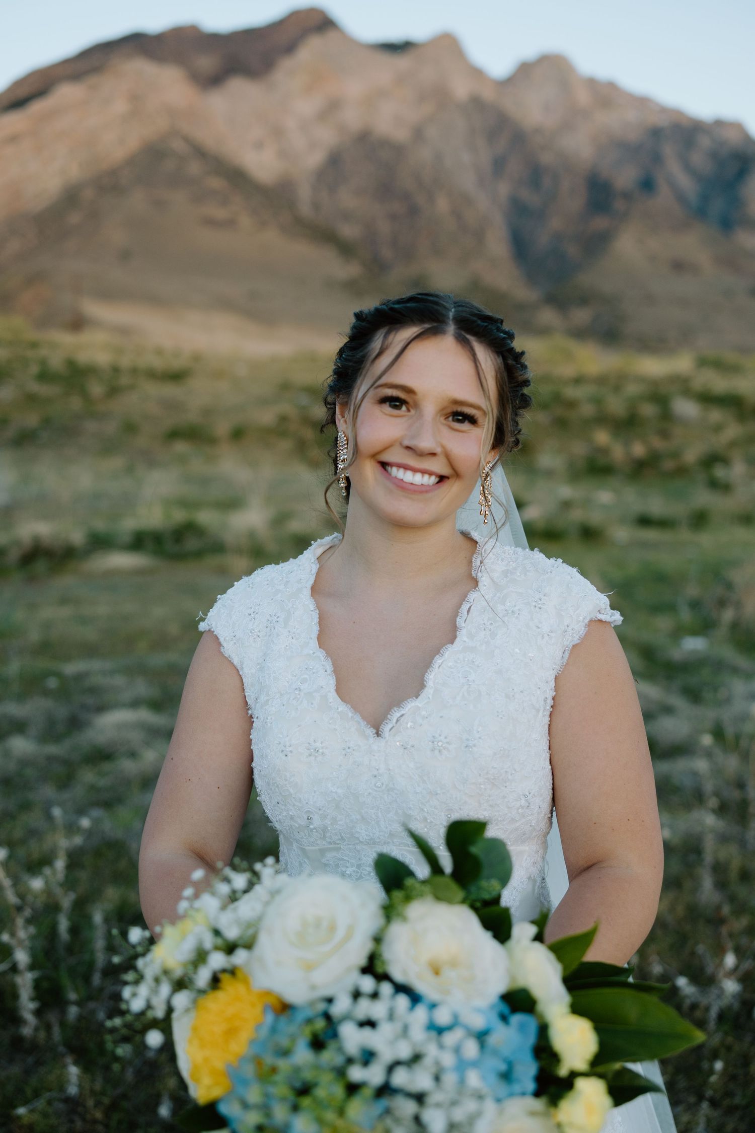 Smiling bride in lace wedding dress holds colorful bouquet with mountain backdrop on her wedding day.