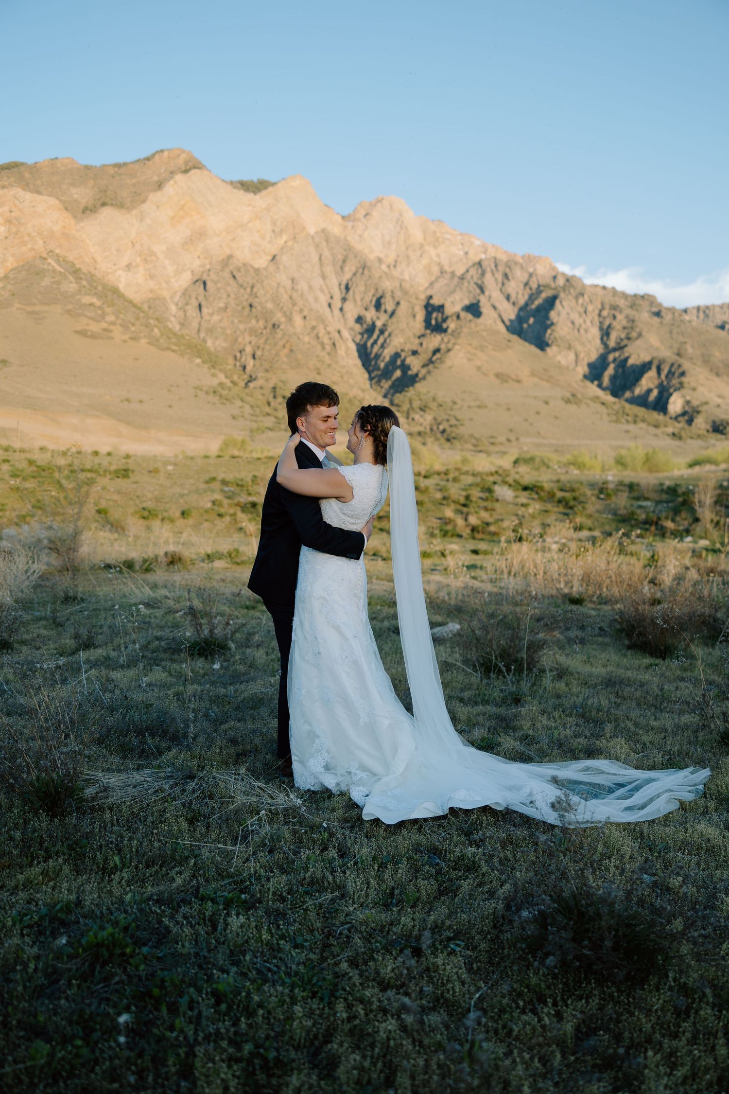 Bride and groom embracing in open mountain field during golden hour sunset light.