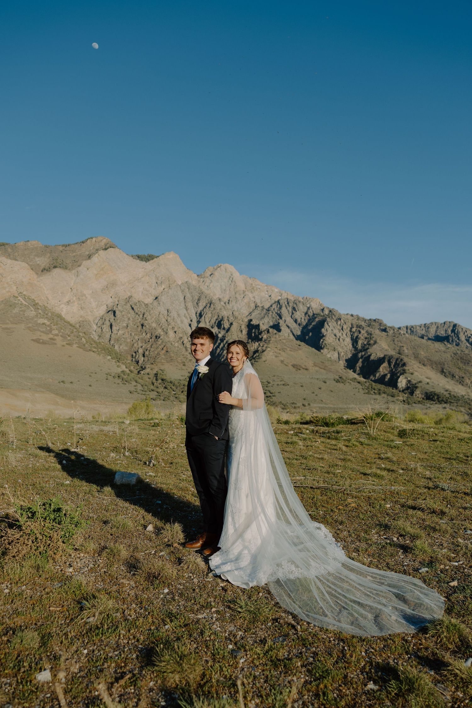 Wedding couple in formal attire pose in a mountain meadow with dramatic rocky peaks and blue sky.