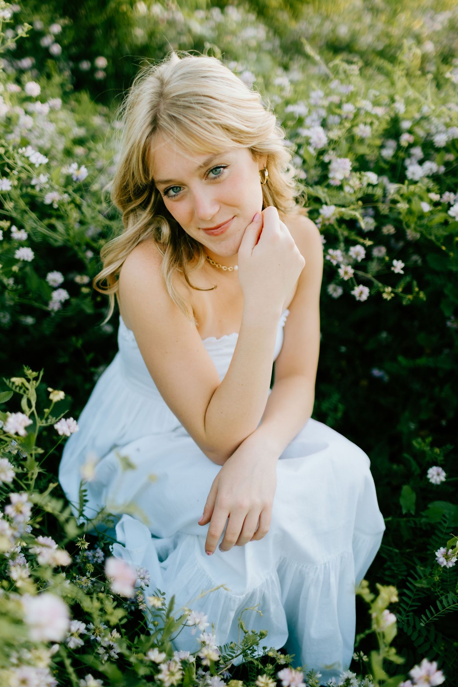 Blonde woman in light blue dress posed among blooming wildflowers, gazing at camera in natural light.